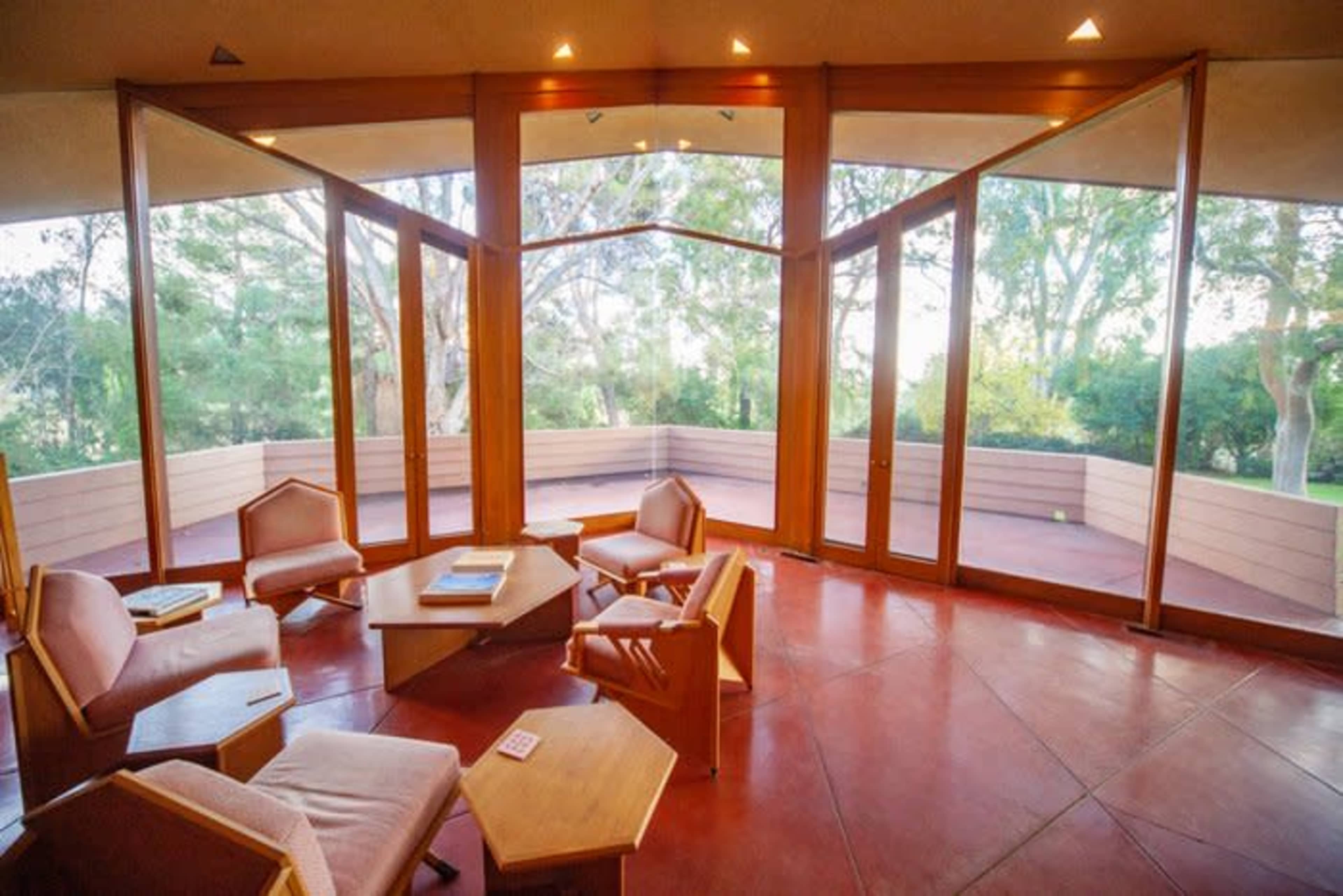 A modern interior space with large glass windows and wooden furniture, opening to an outdoor area surrounded by trees.