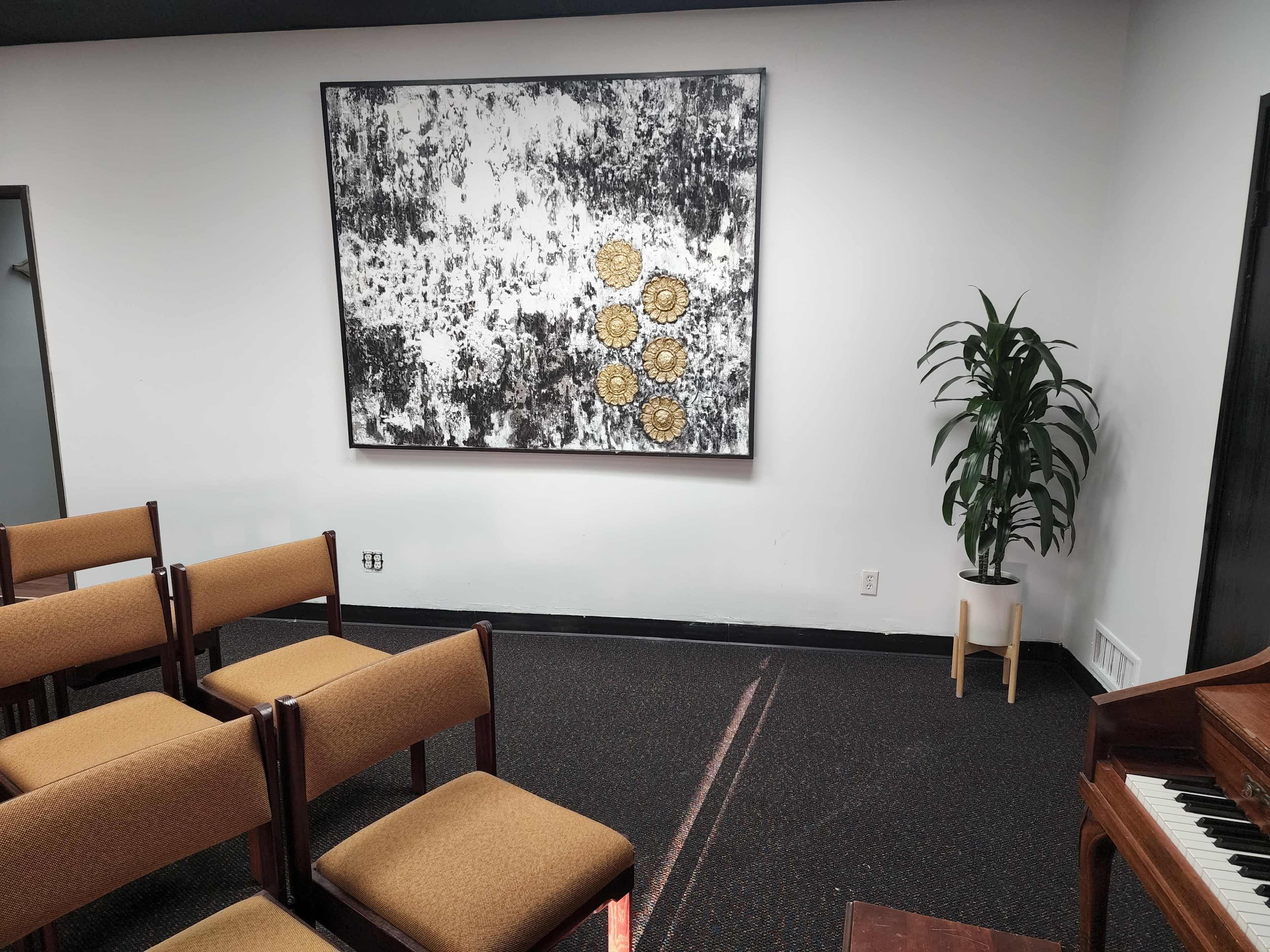 A small room with brown chairs arranged in front of an abstract artwork featuring gold circles on a textured background, a potted plant in the corner, and a piano nearby.