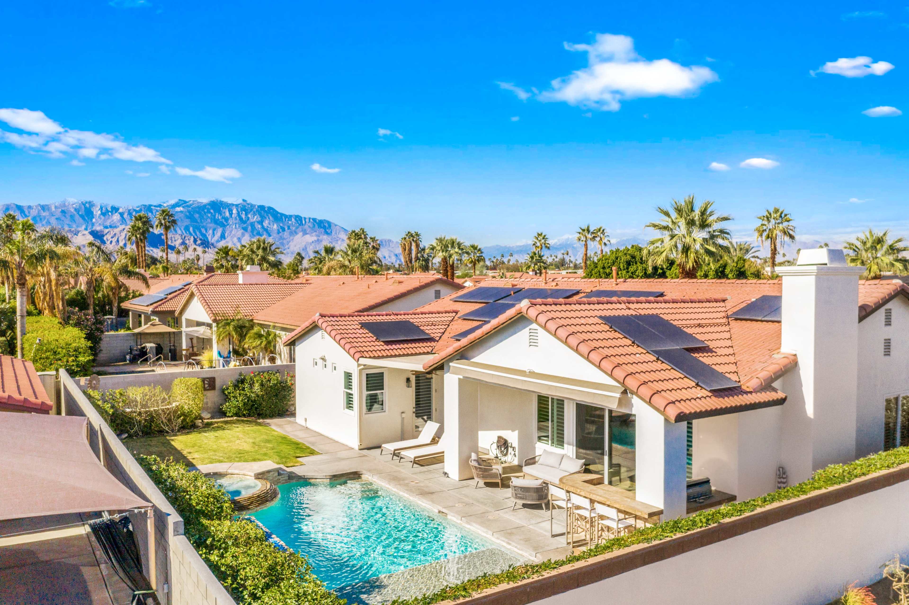 A modern house with a swimming pool and solar panels is surrounded by palm trees, with mountains visible in the background.
