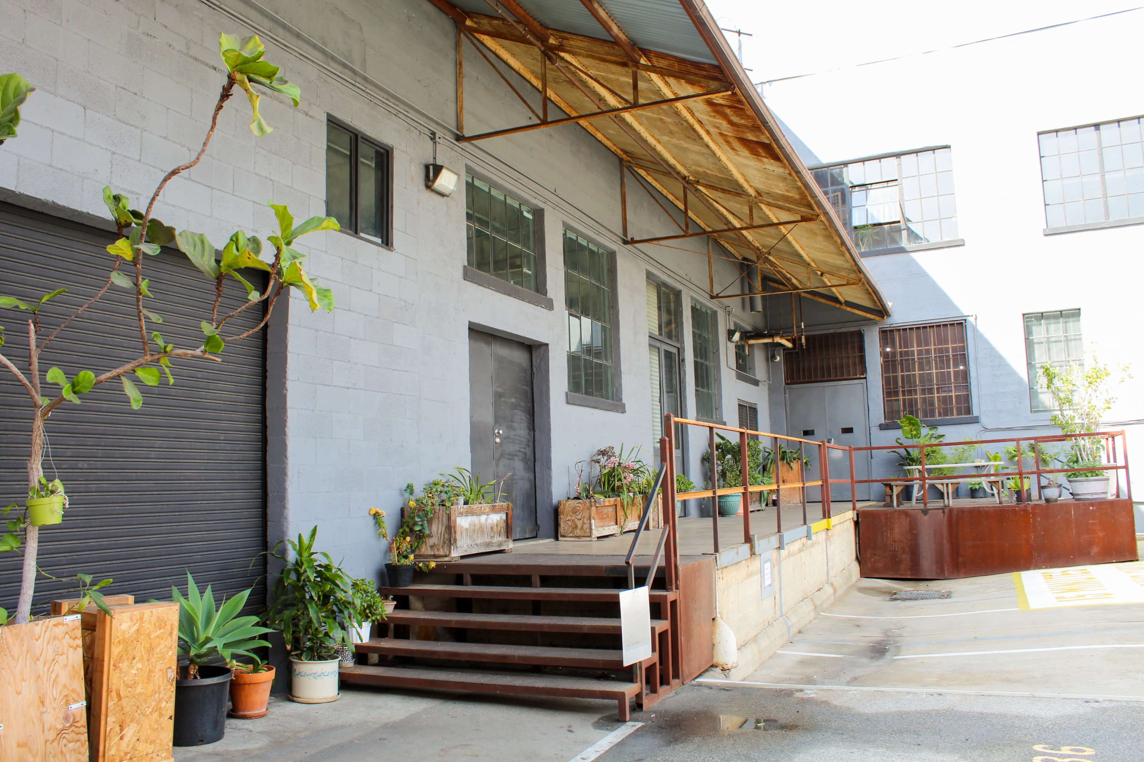 The image shows a gray industrial building with a wooden awning, steps leading up to a door, and various plants in containers along the entrance.