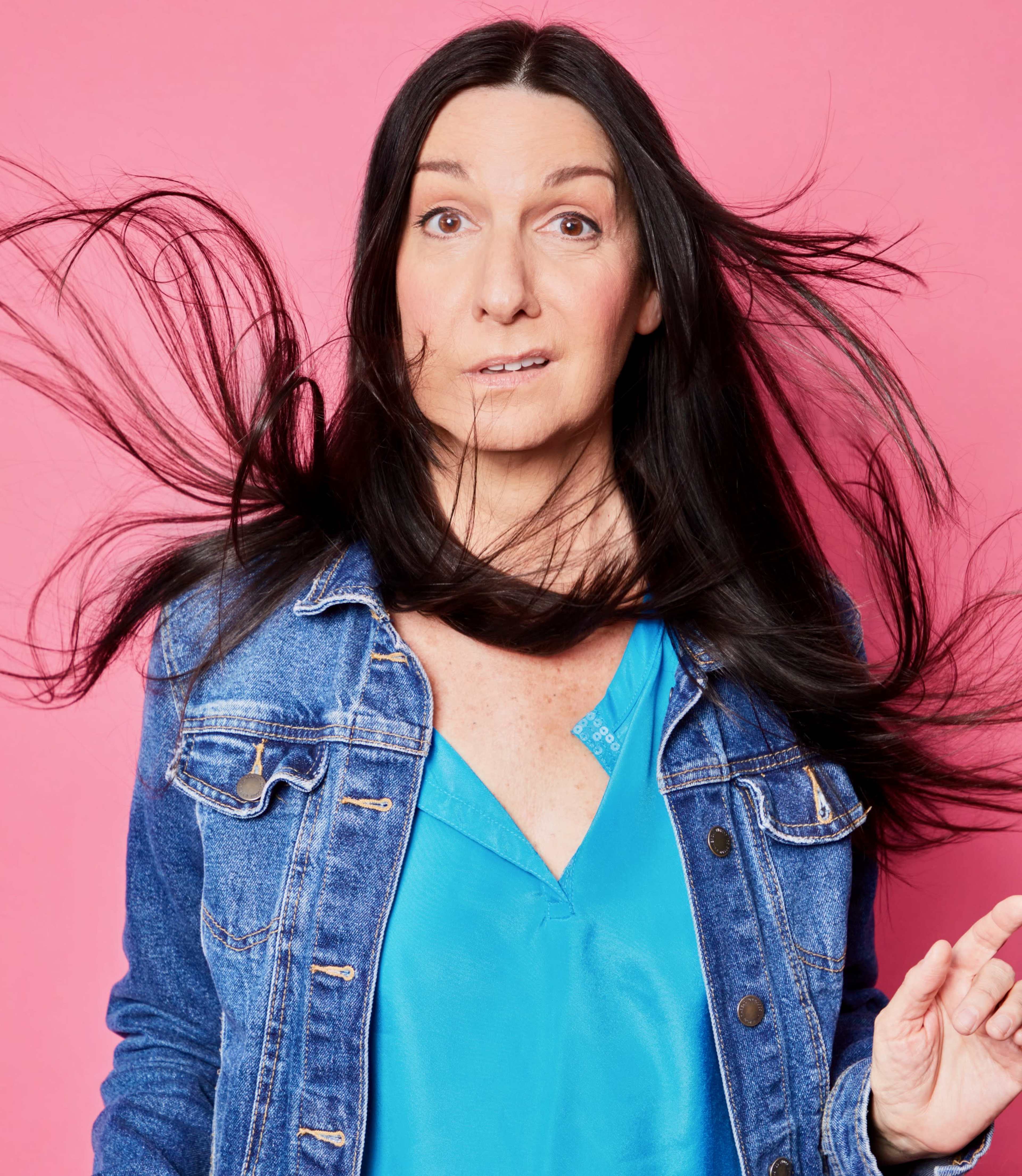 A woman with long, flowing hair stands against a pink background, wearing a blue top and a denim jacket.