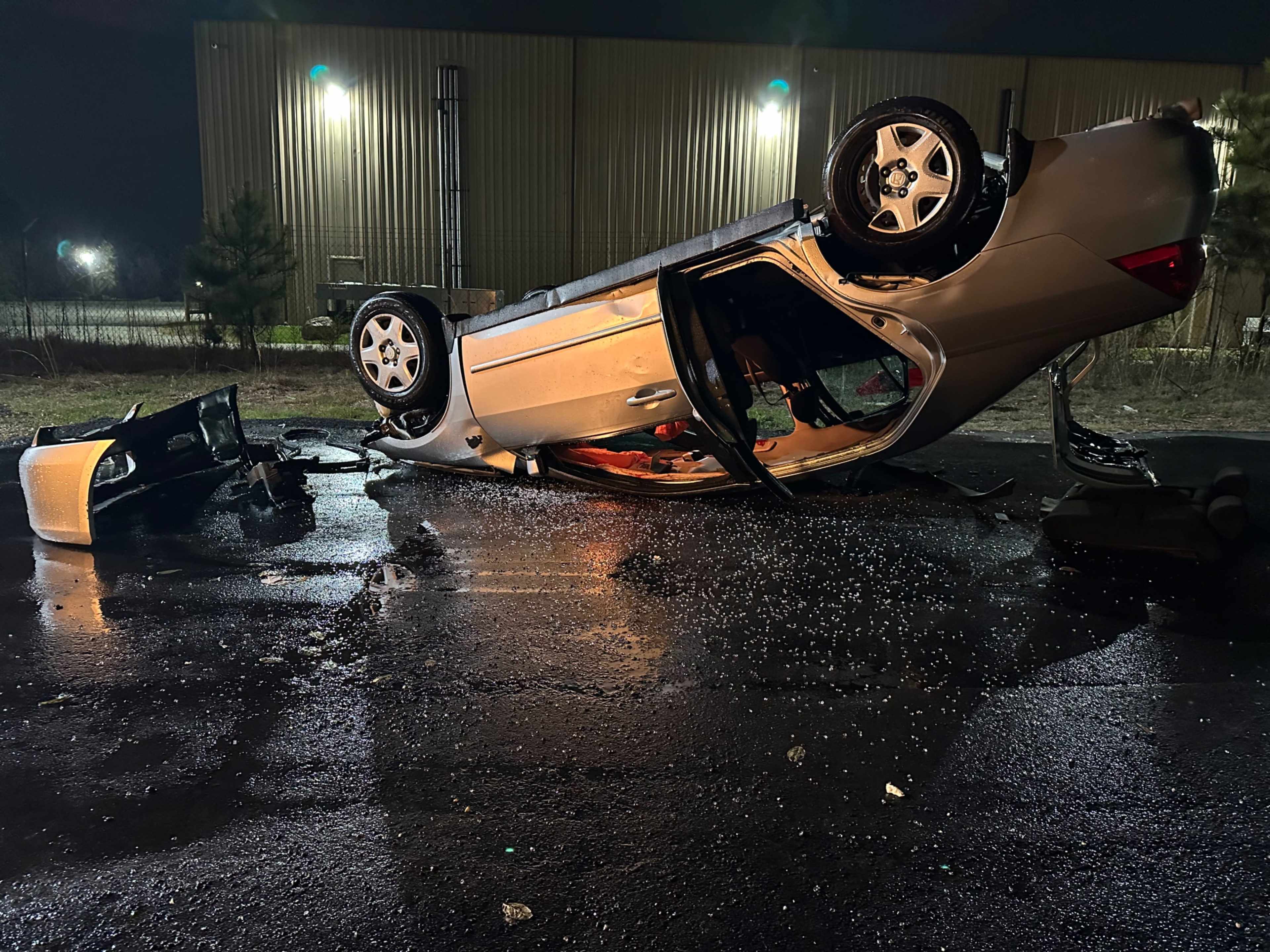 A silver car is overturned on its roof on a dark, wet roadway, with debris scattered nearby.
