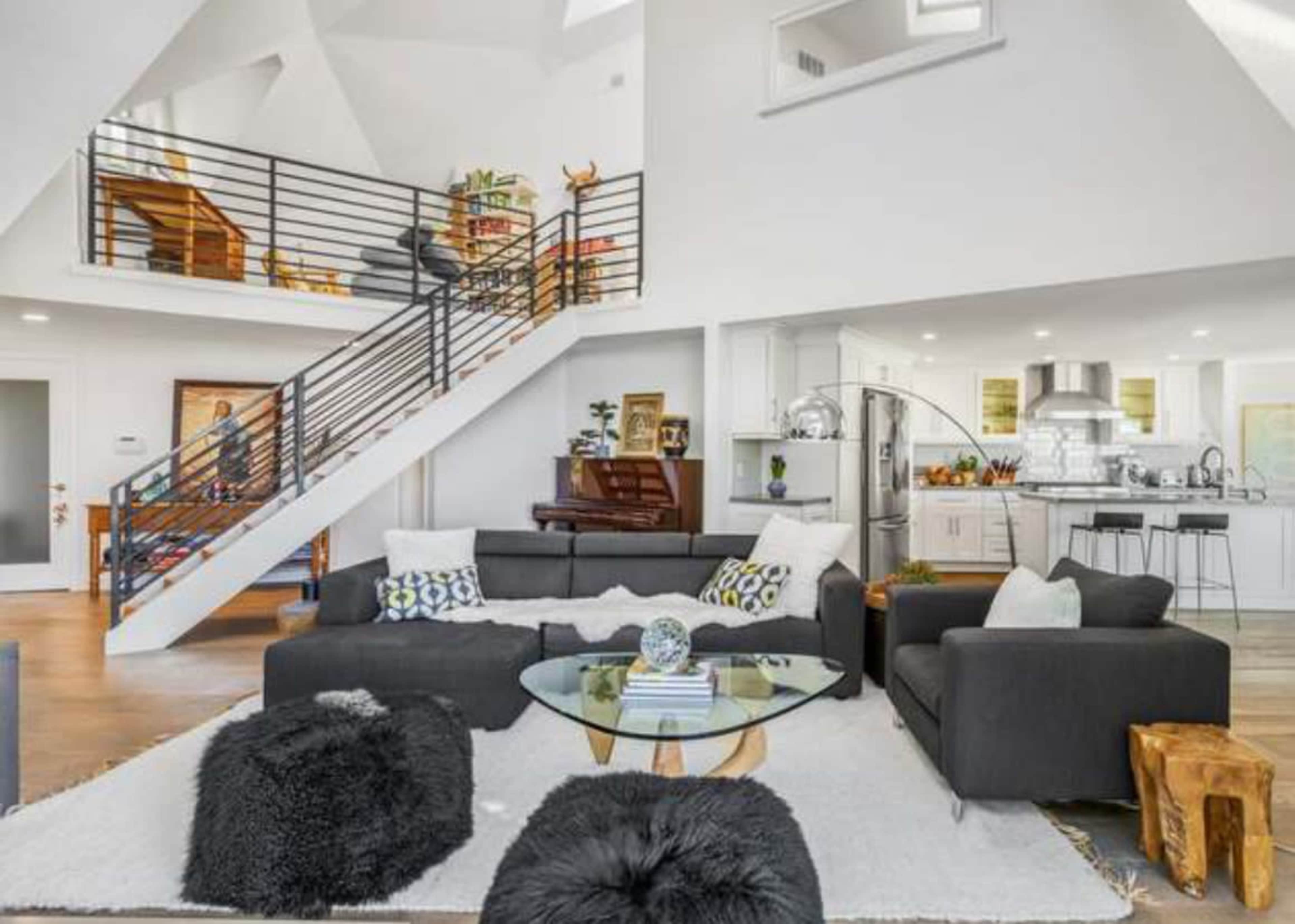 The image shows a modern living room with a gray sofa, a glass coffee table, and a staircase leading to an upper floor, adjacent to an open kitchen with a piano and dining area.