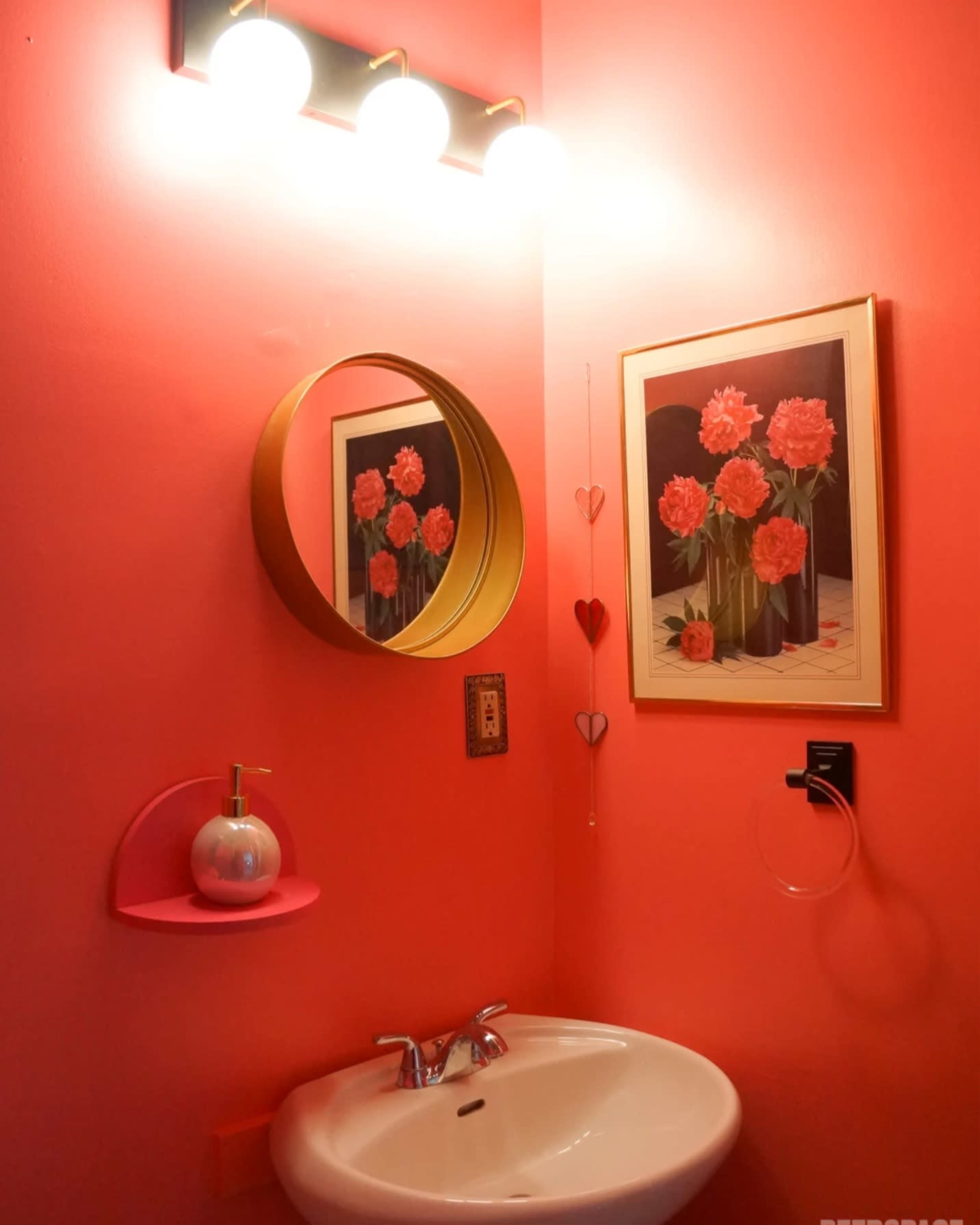 A small bathroom with pink walls, a circular mirror, a framed floral painting, and a white basin with a soap dispenser on a pink shelf.