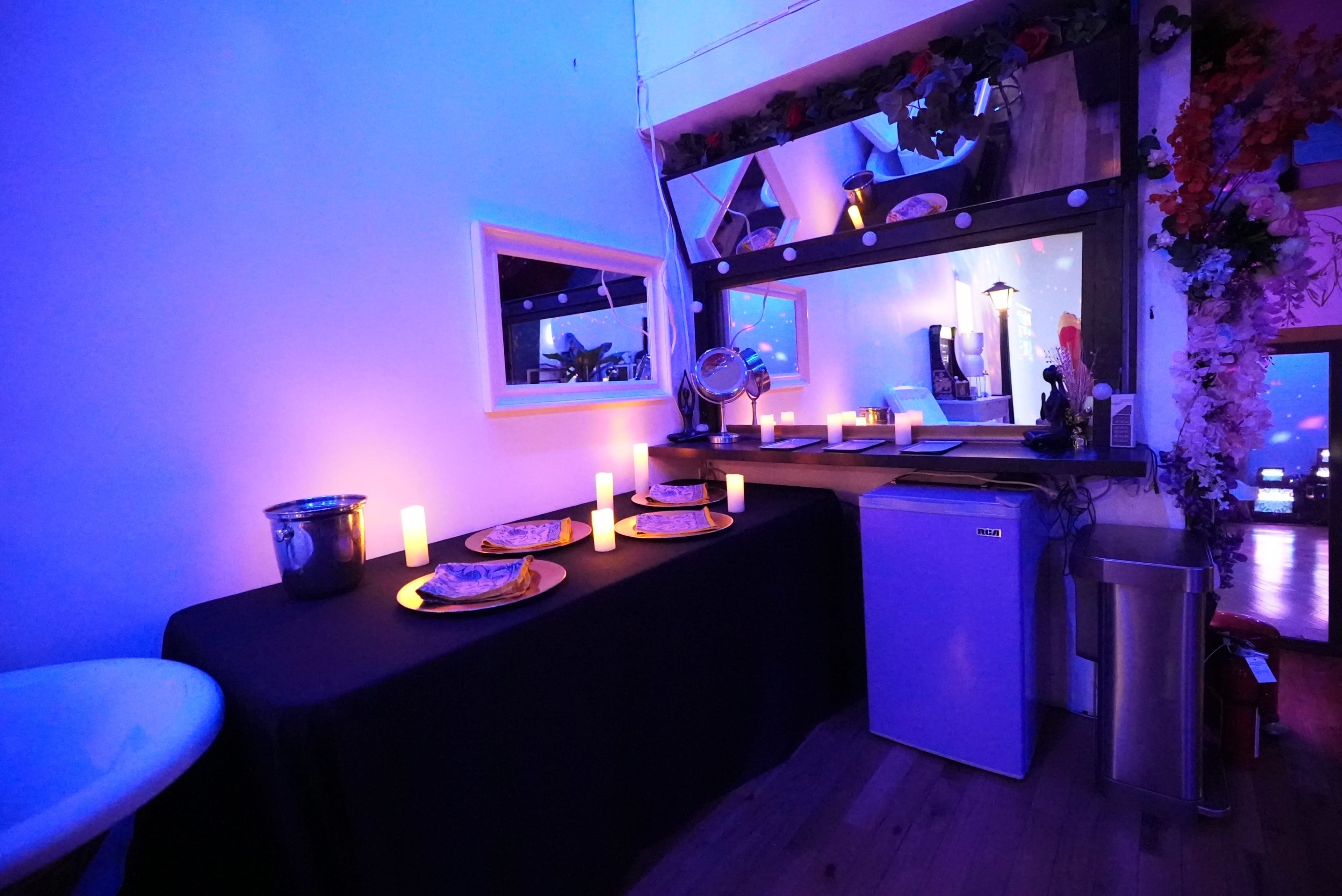 The image shows a dimly lit room with a black table covered with plates and candles, featuring a mirror and a small fridge nearby.
