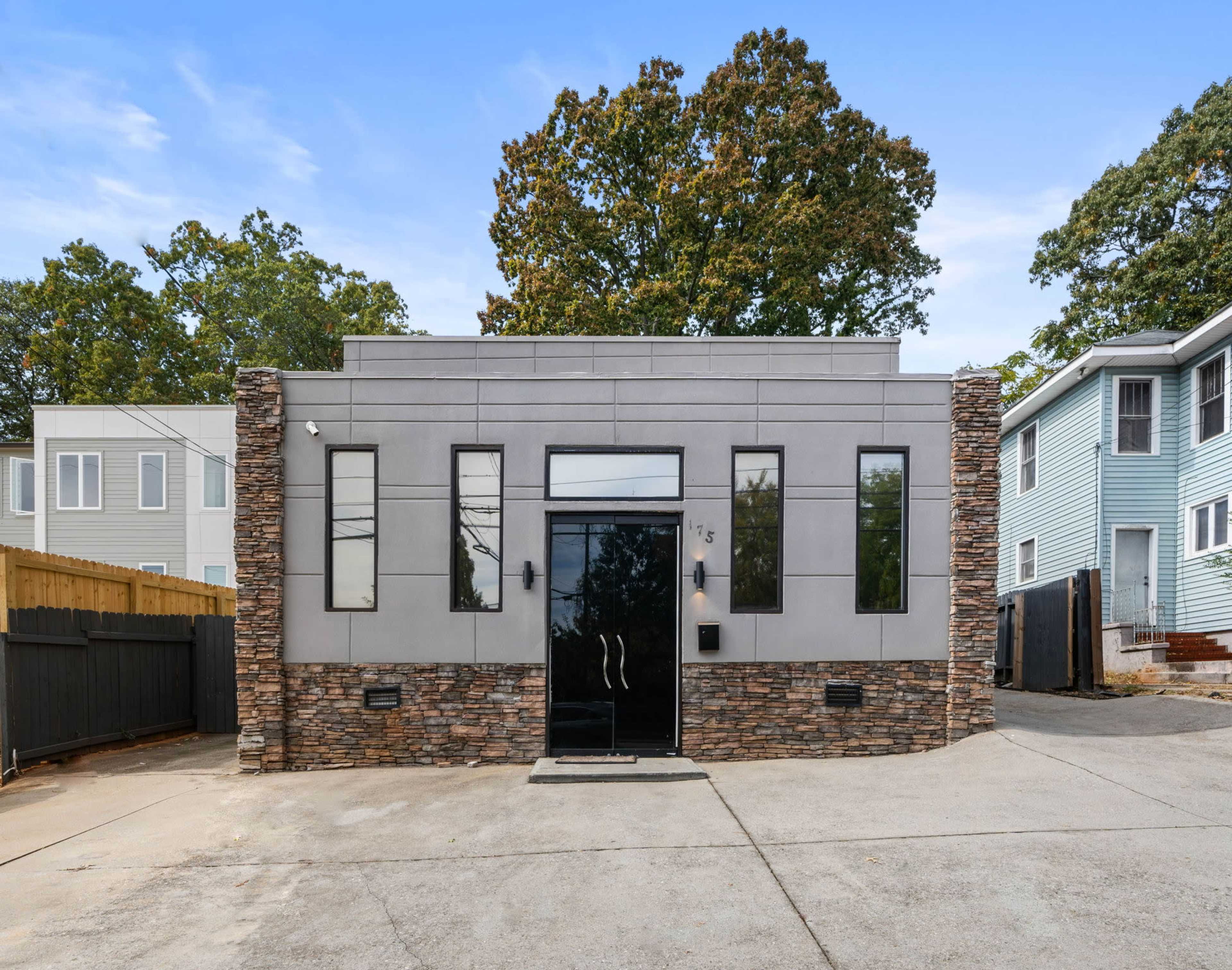 The image shows a modern gray building with a black front door and stone accents, surrounded by a paved area and residential homes in the background.