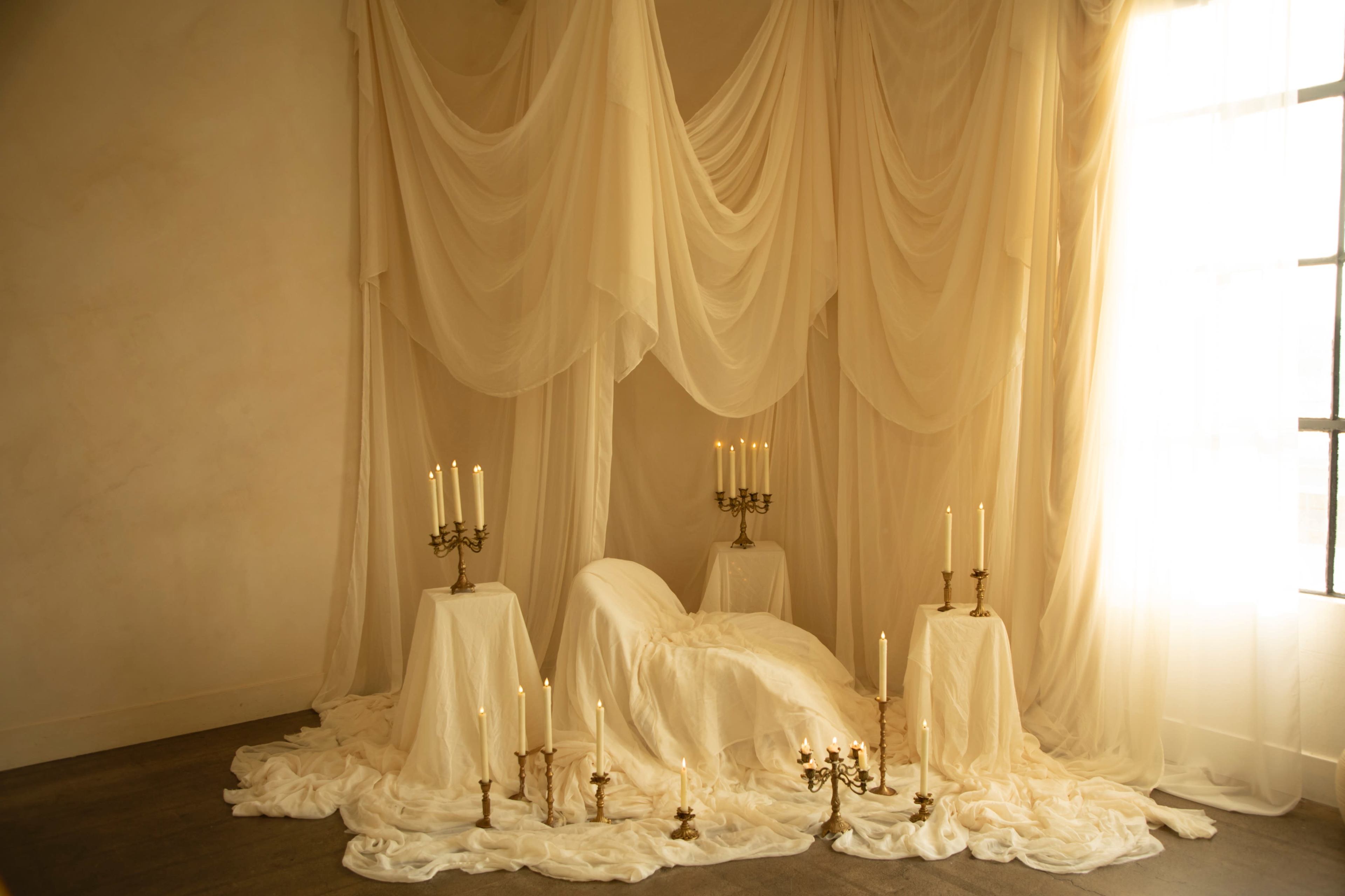 The scene features a softly lit room adorned with flowing sheer fabric and candles arranged around a draped bed.