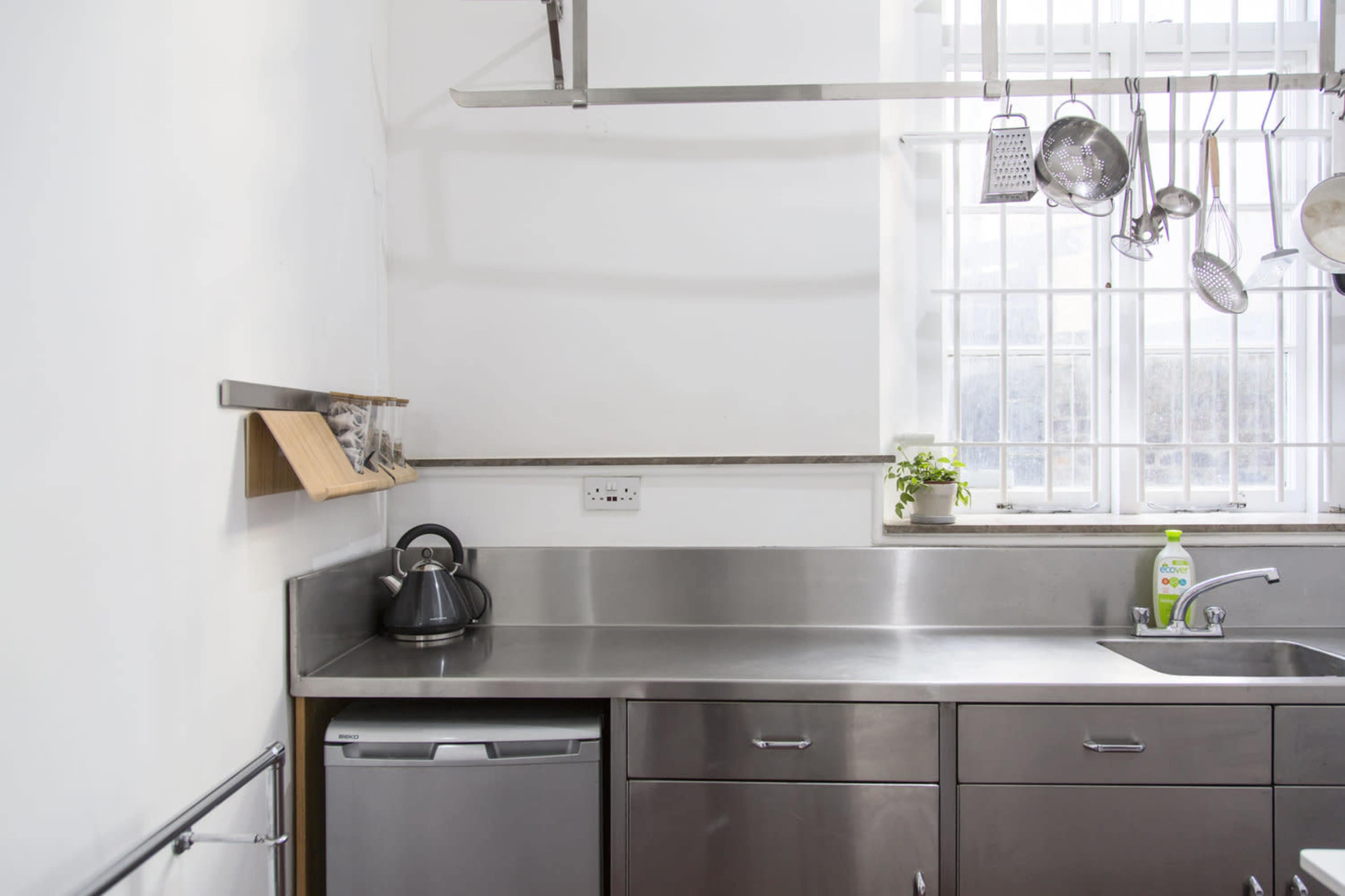 A modern kitchen with stainless steel countertops, a kettle on the side, a small fridge, and hanging kitchen utensils.