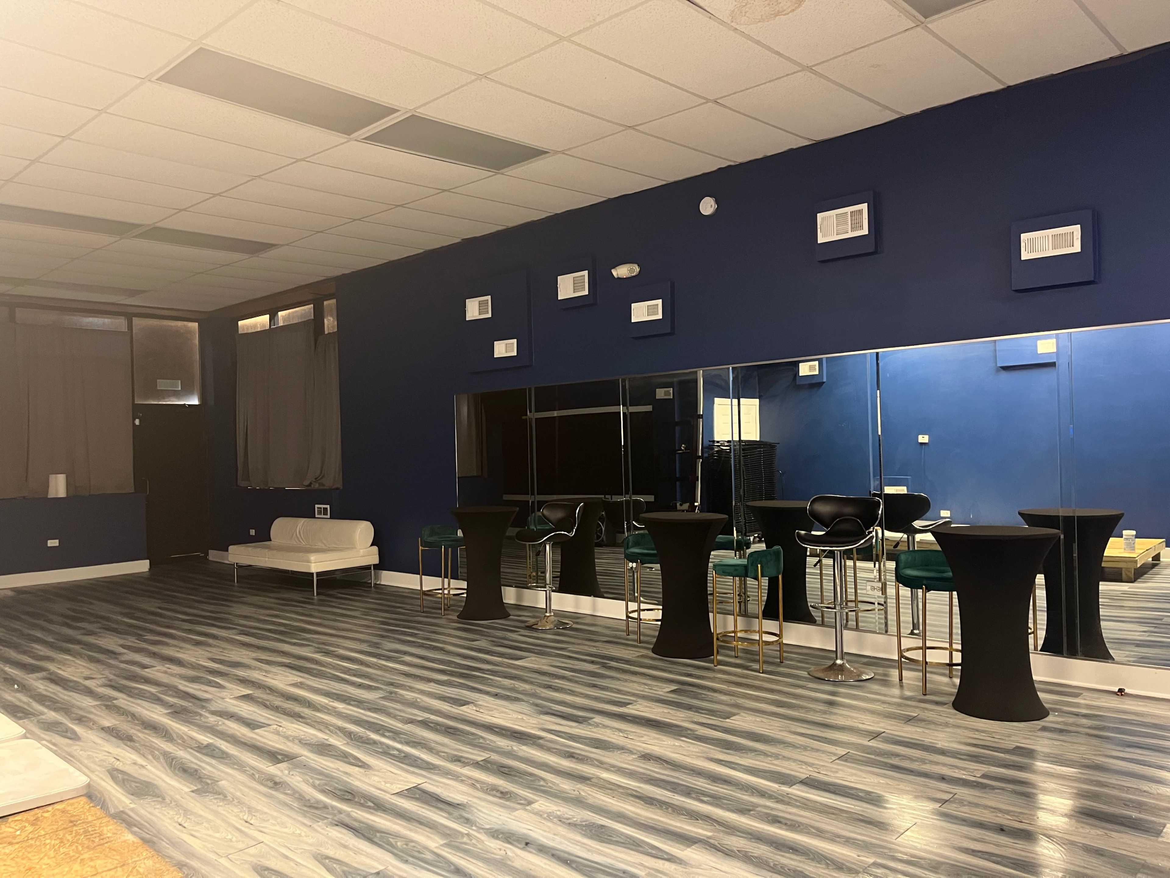 A spacious room with blue walls, wooden flooring, several tall black tables, and a large mirror reflecting the seating area.