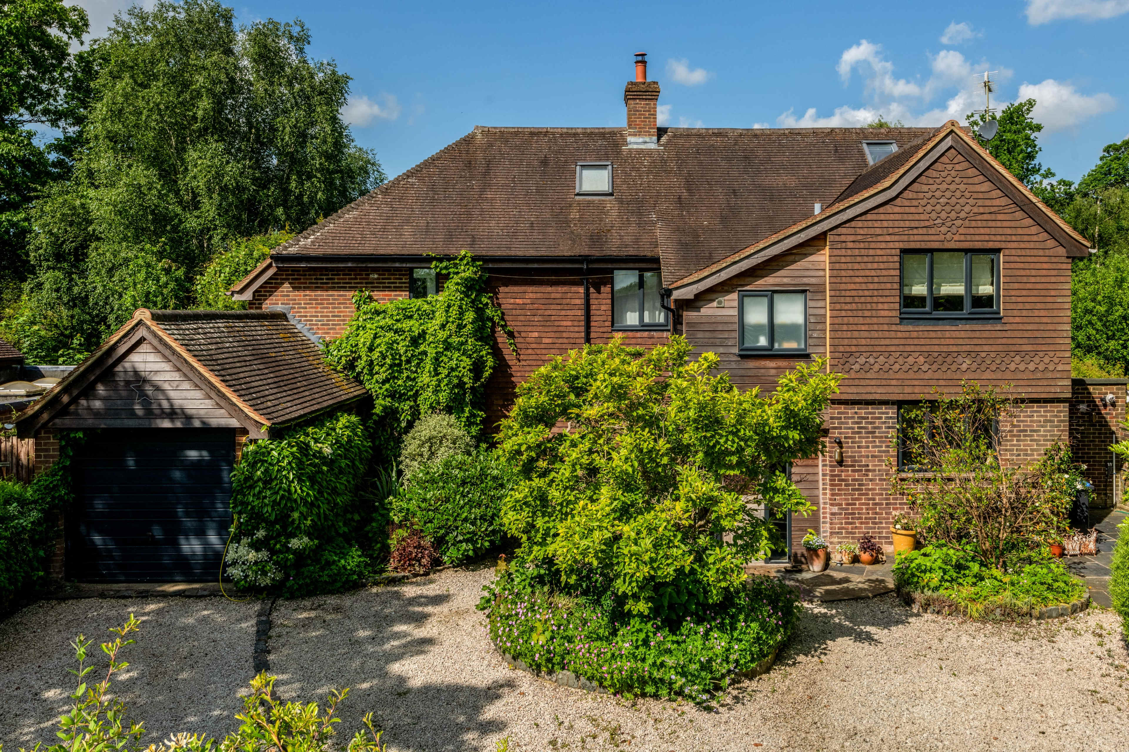 A two-story brick house with a gravel driveway is surrounded by greenery and landscaped gardens.