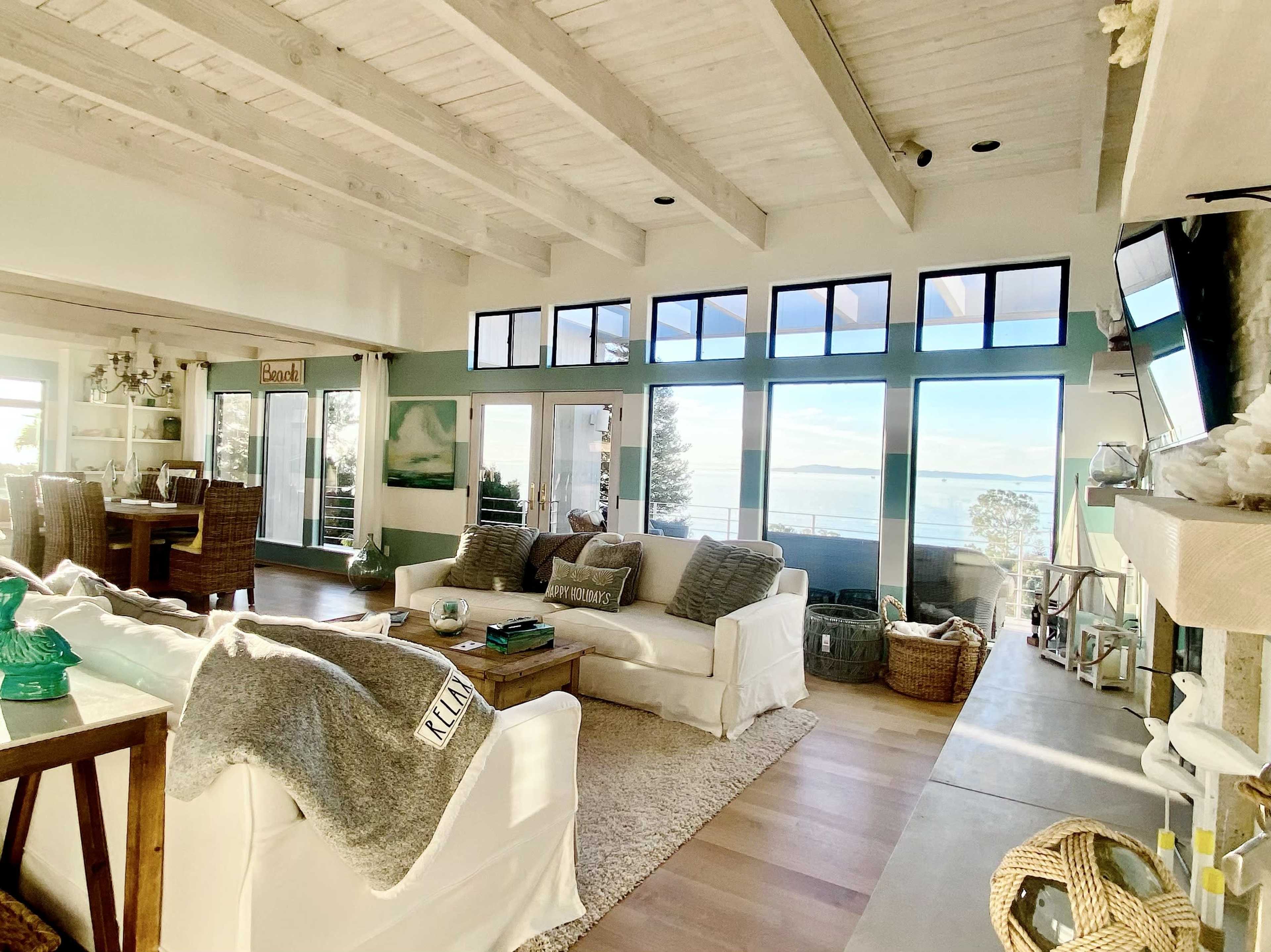 A bright living room features a cozy seating area, large windows with a view of water, and a dining space in the background.