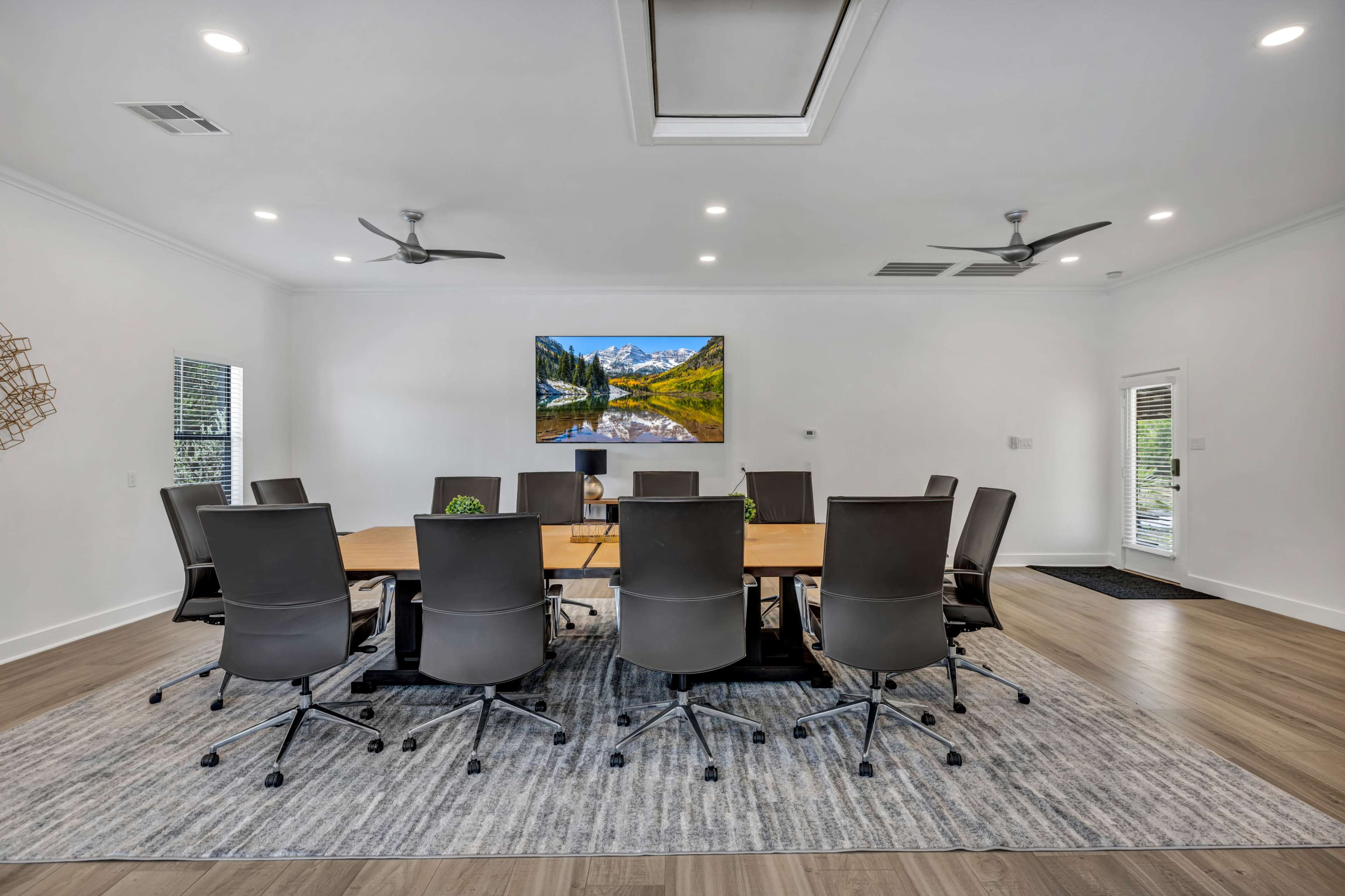A modern conference room features a large wooden table surrounded by eight black ergonomic chairs, with a landscape painting on the wall and natural light coming through the windows.