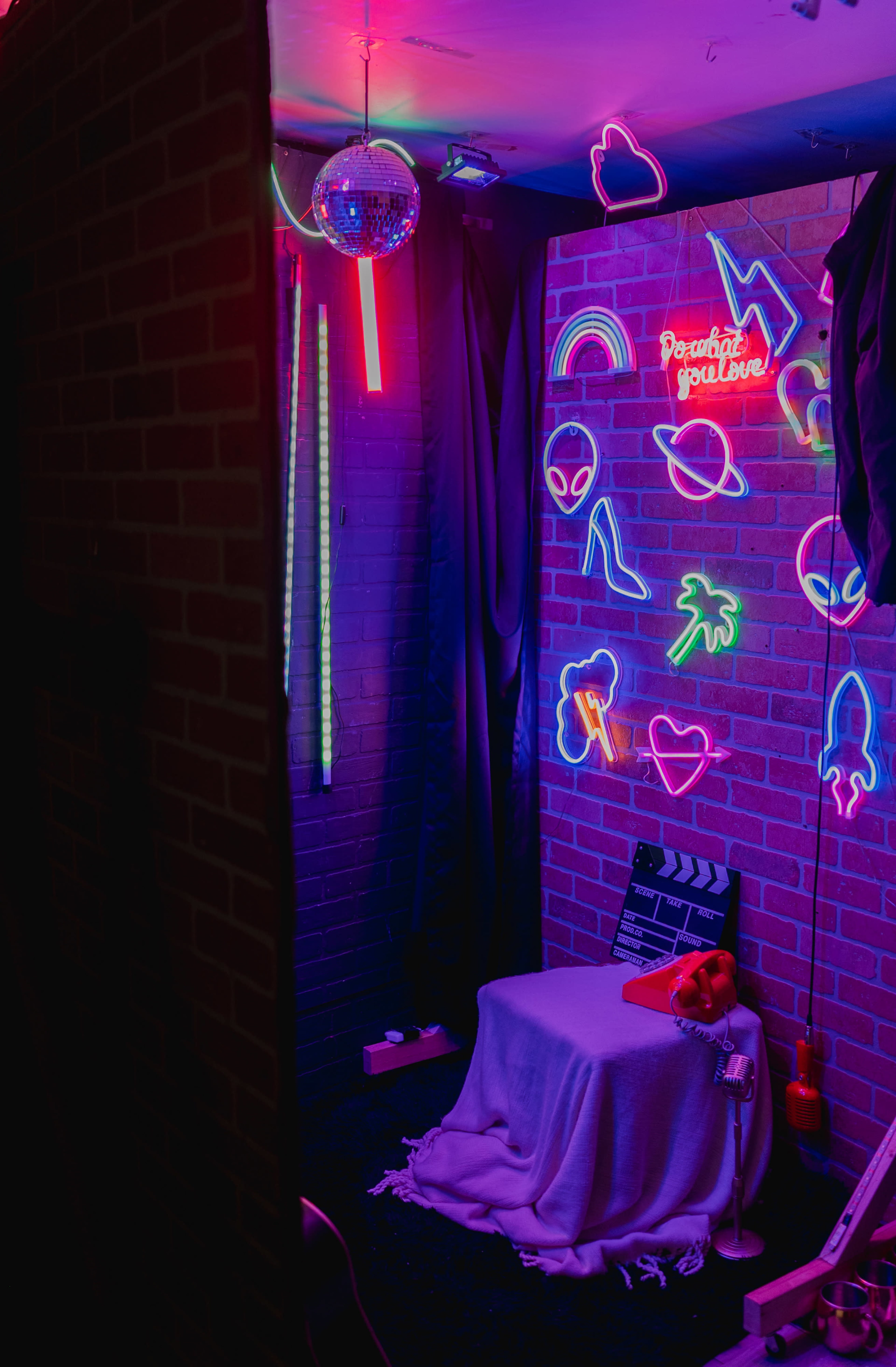 A dimly lit room featuring a wall adorned with neon signs, a disco ball, and a chair covered with a white cloth in front of a brick wall.