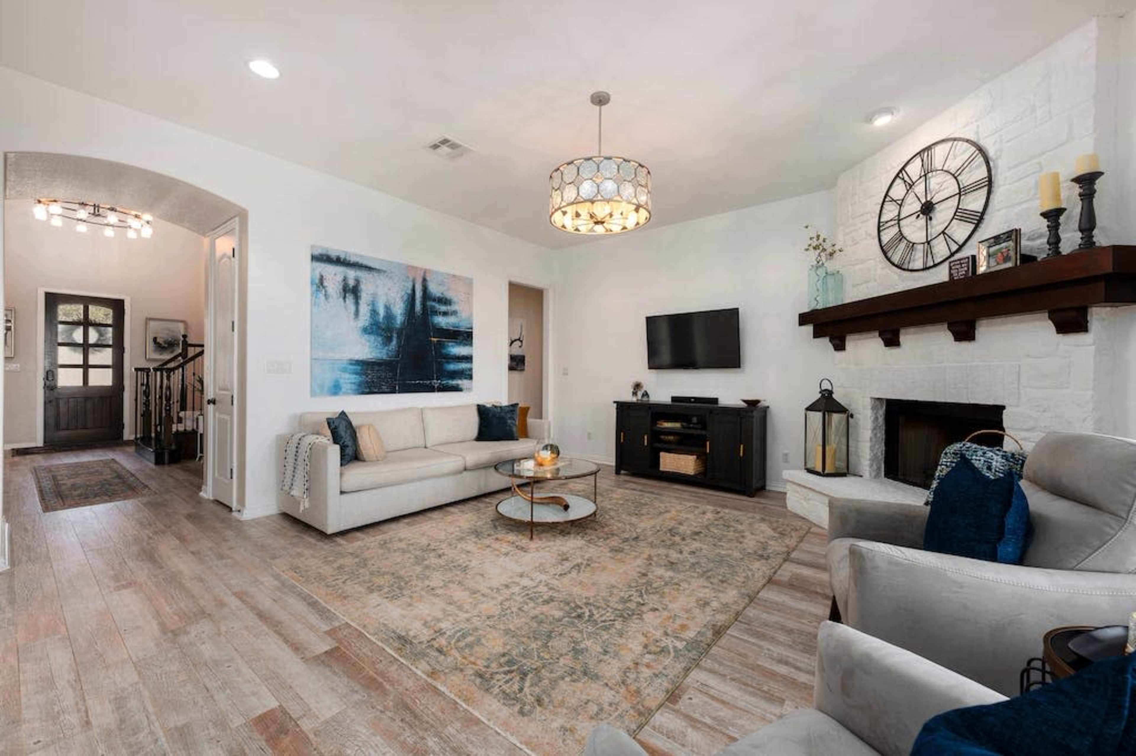 The living room features a white sofa, a round coffee table, a dark media console, and a stone fireplace, with a large wall art piece and a decorative chandelier overhead.