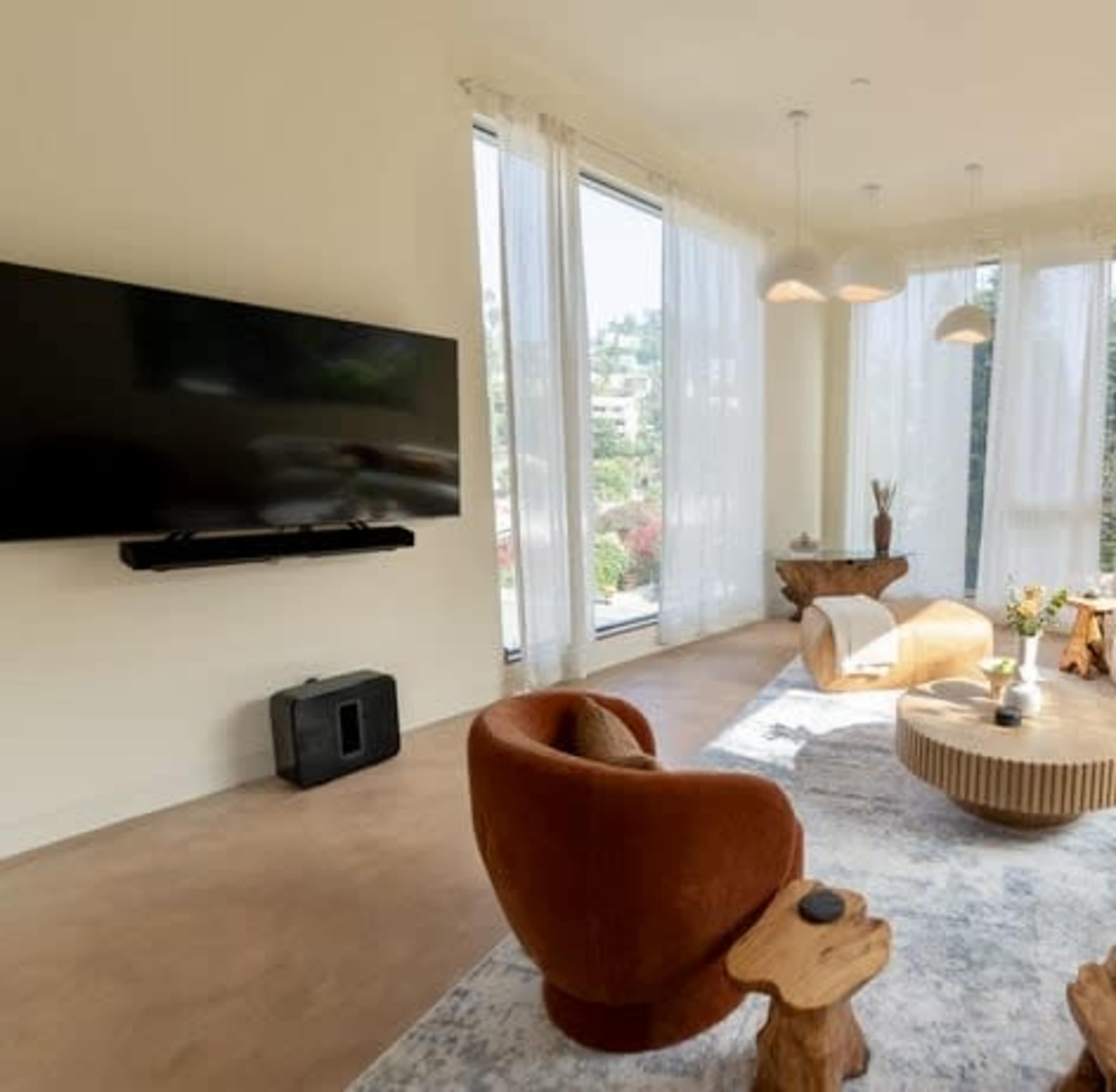 A modern living room features large windows, a flat-screen TV mounted on the wall, and a round wooden coffee table surrounded by two armchairs and natural wood side tables.