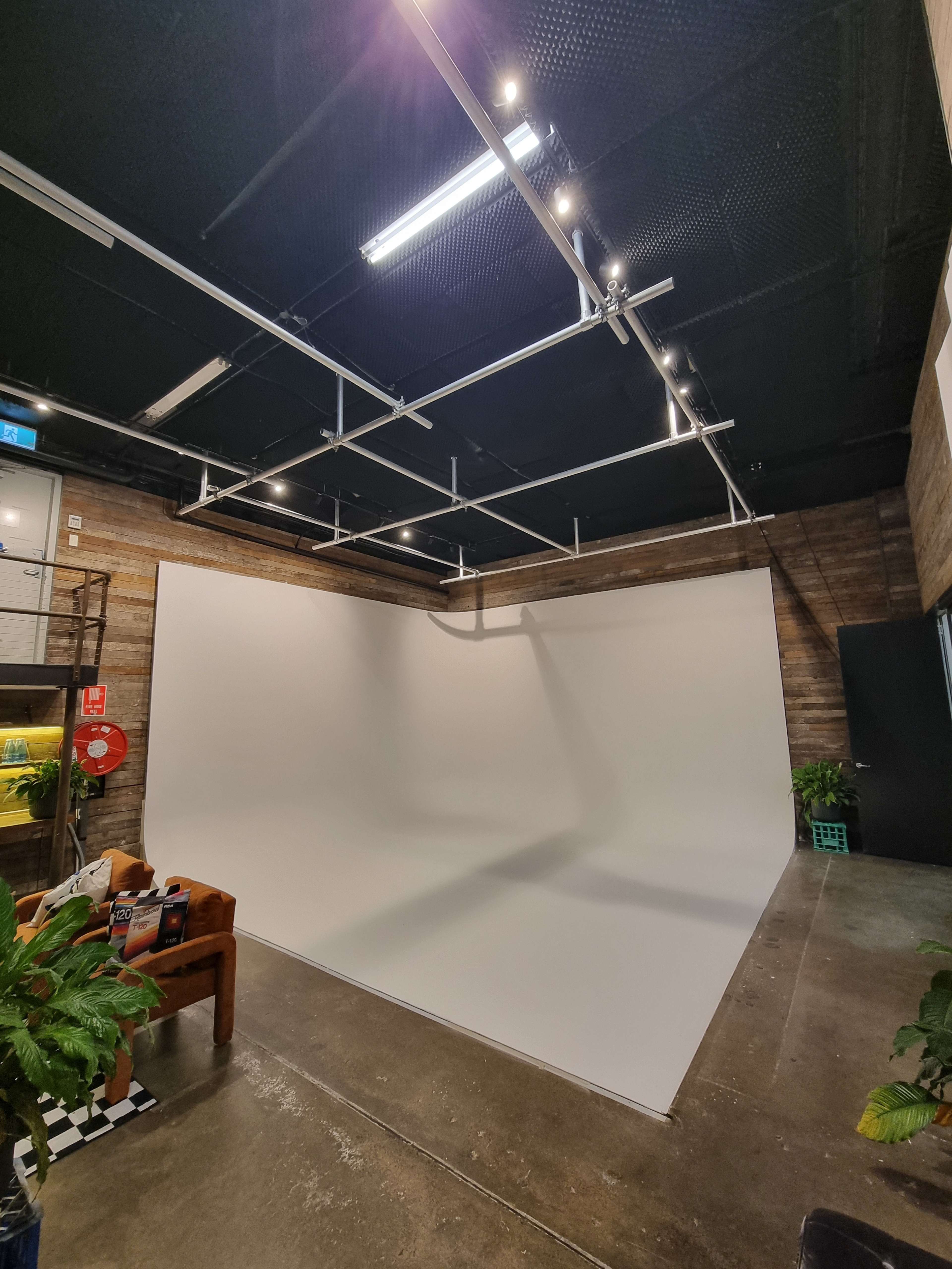 The image shows a spacious photography studio featuring a large white backdrop and overhead lighting, with a seating area in the corner.