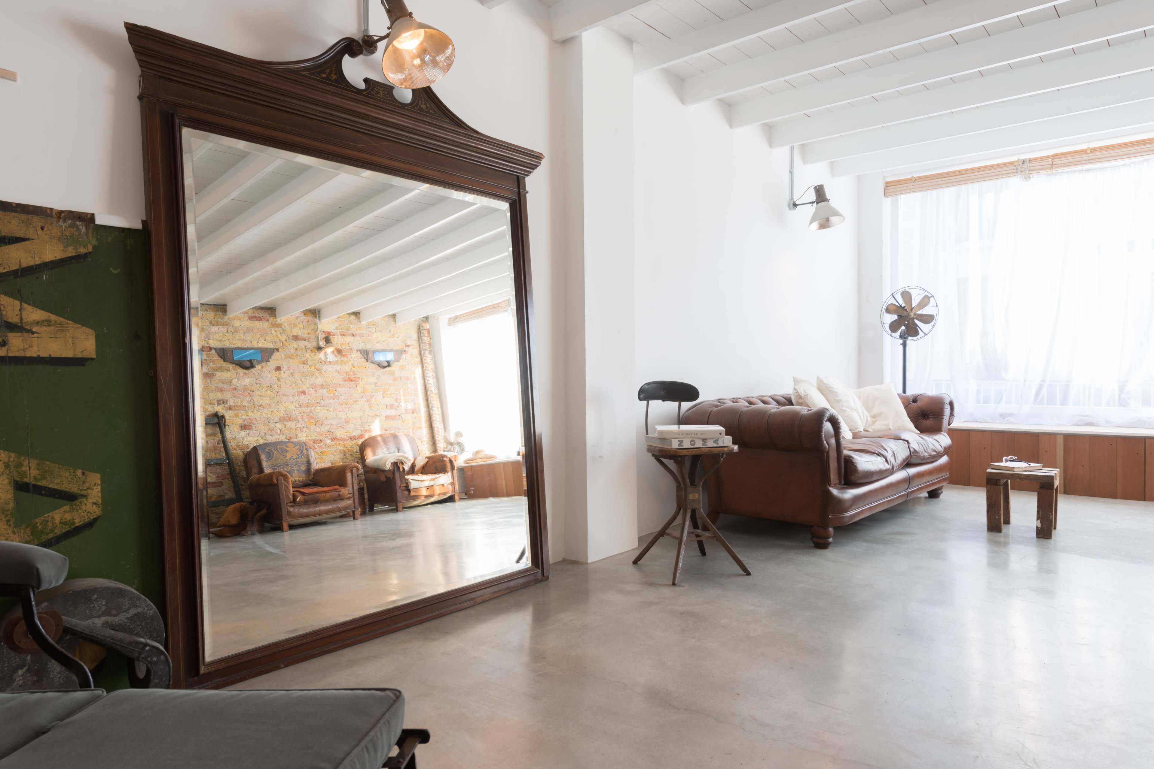A large ornate mirror reflects a bright room with a brown leather sofa, wooden furniture, and exposed brick walls.