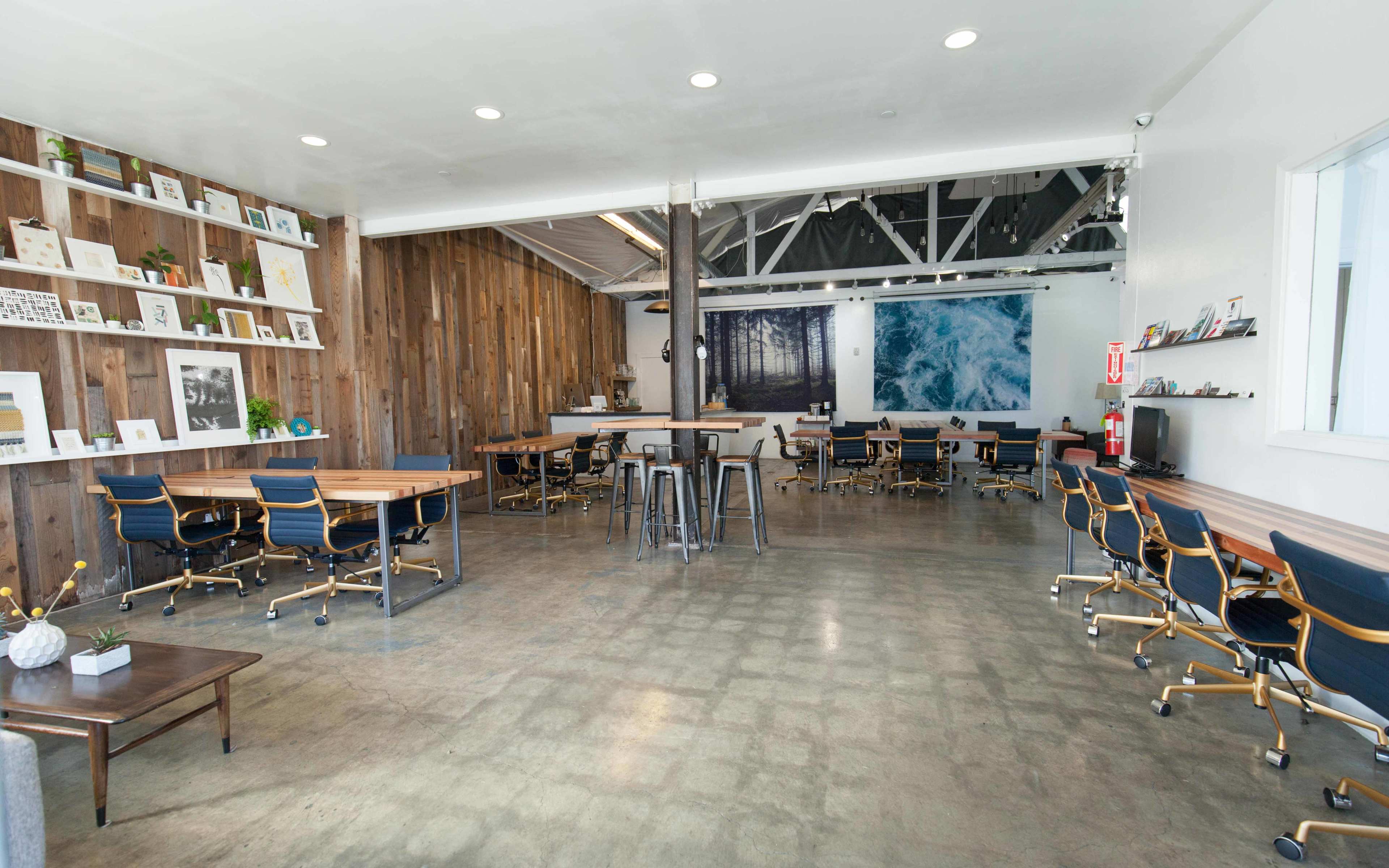 A spacious, modern workspace featuring wooden walls, multiple tables with chairs, and a large artwork of ocean waves.