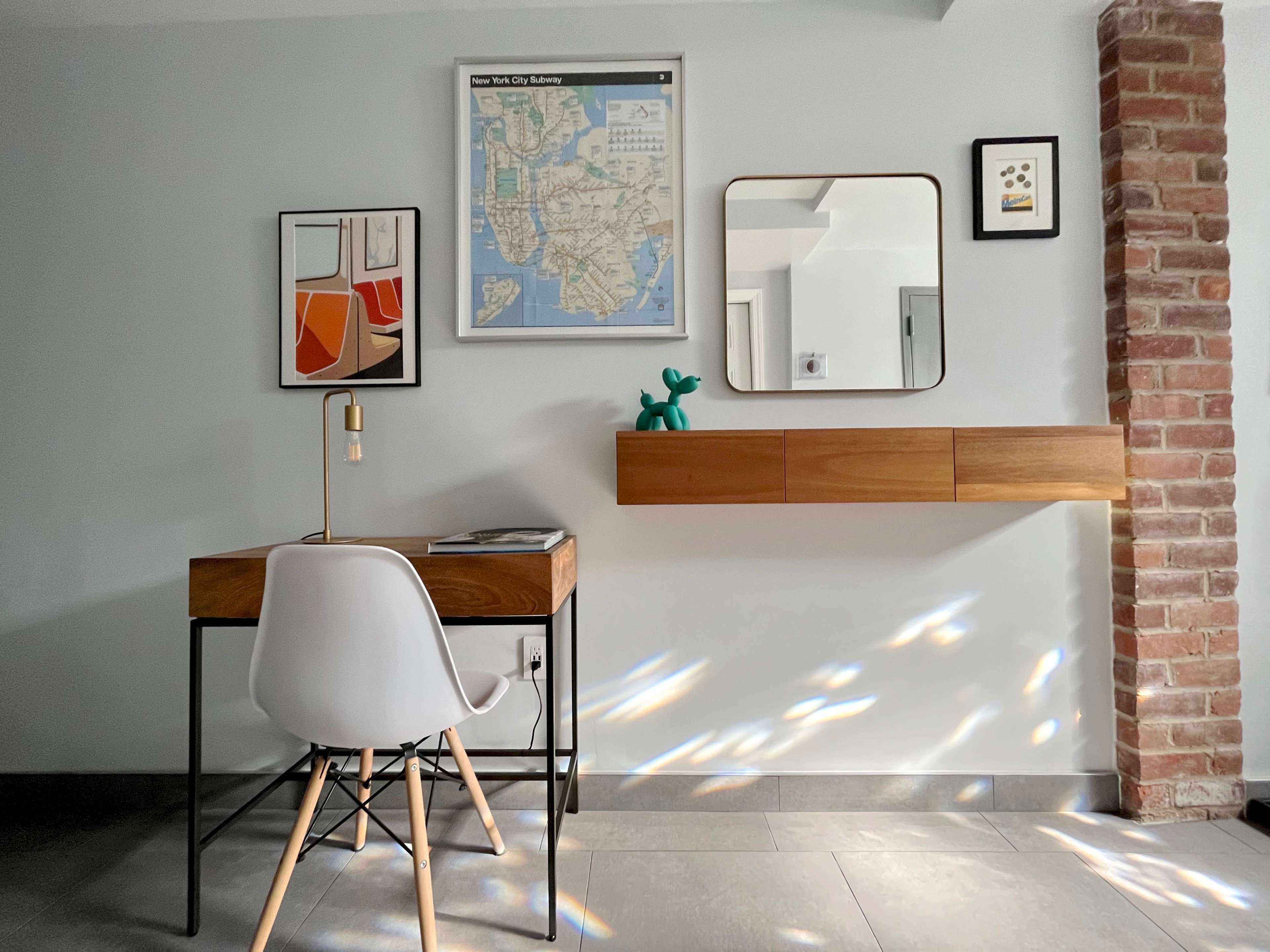 A desk with a chair is set against a wall adorned with framed artwork and a map, next to a small, mounted shelf.
