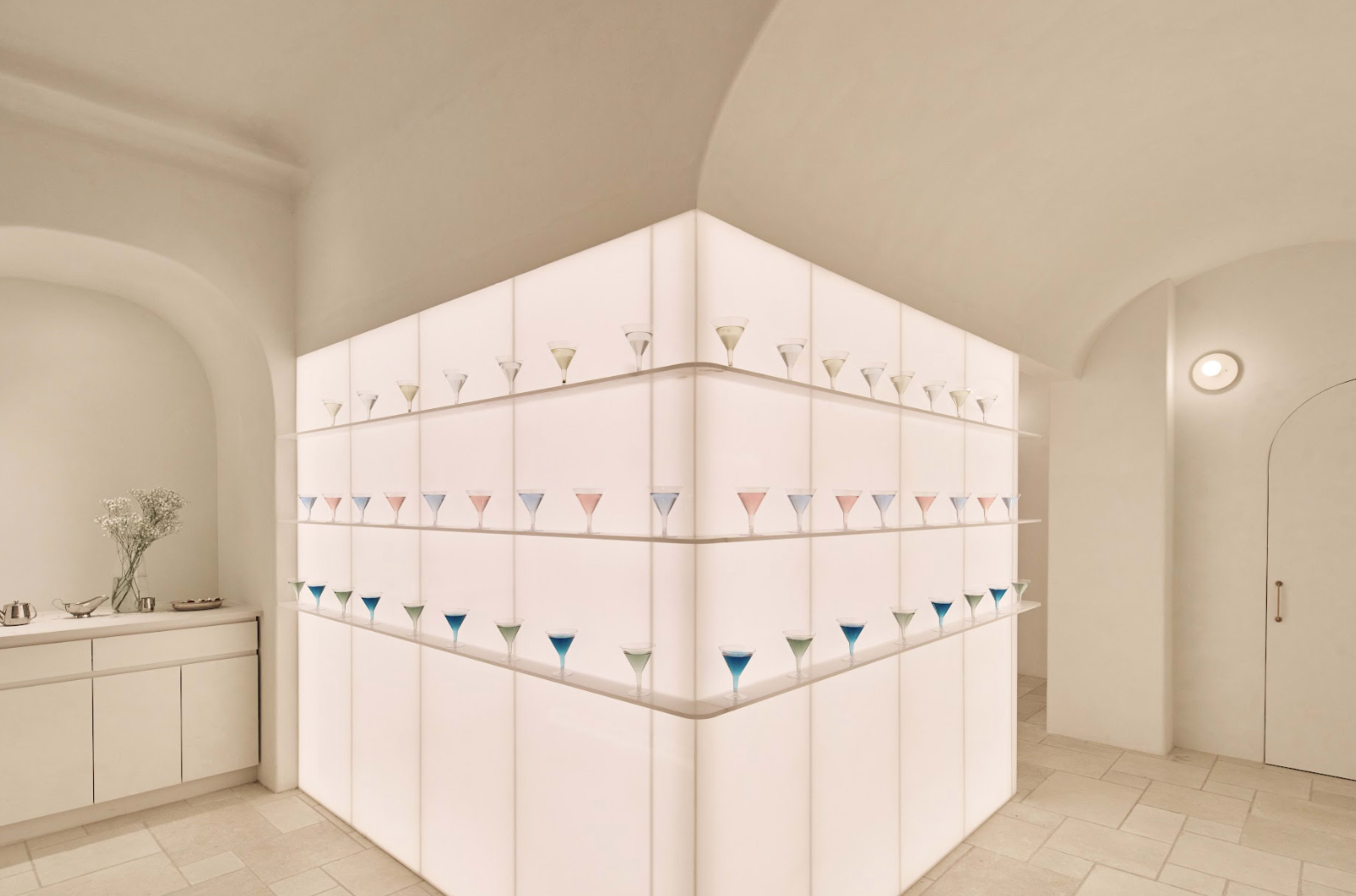The image shows a modern, well-lit interior with shelves displaying numerous colorful glass cups arranged in a geometric pattern.