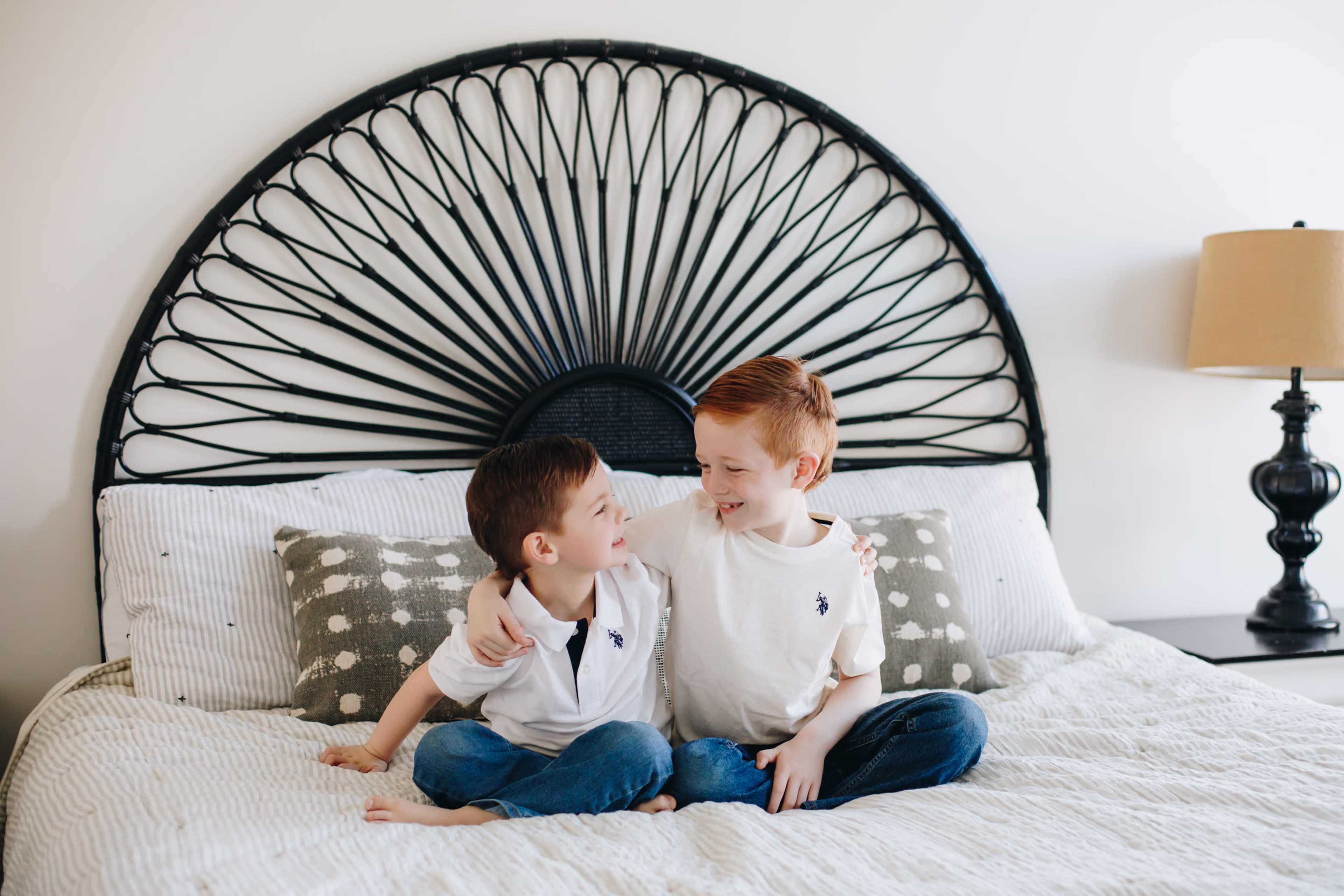 Two young boys sit on a bed with a large decorative headboard, smiling and embracing each other.