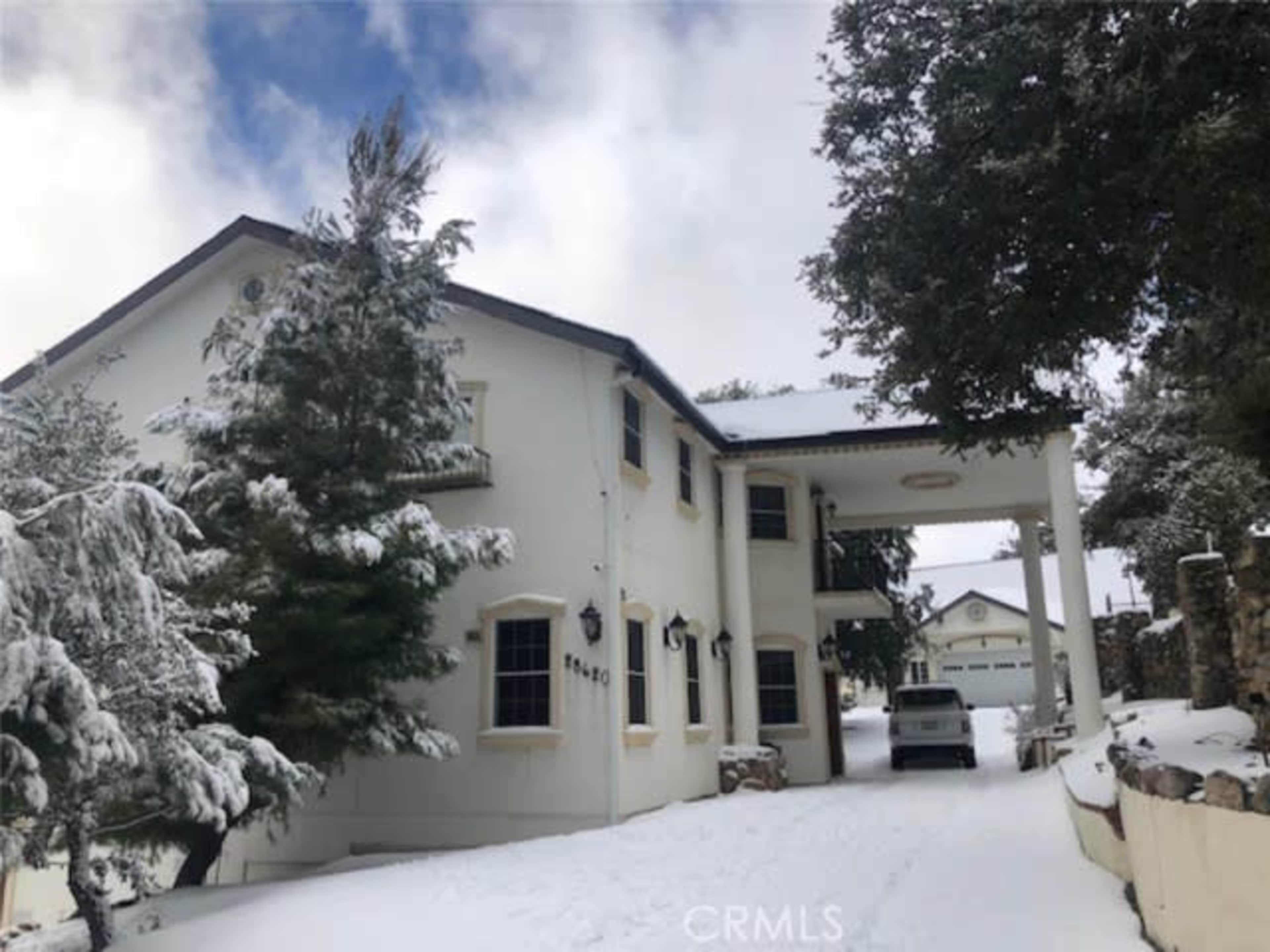 A large white two-story house is surrounded by snow-covered trees, with a vehicle parked in the driveway.