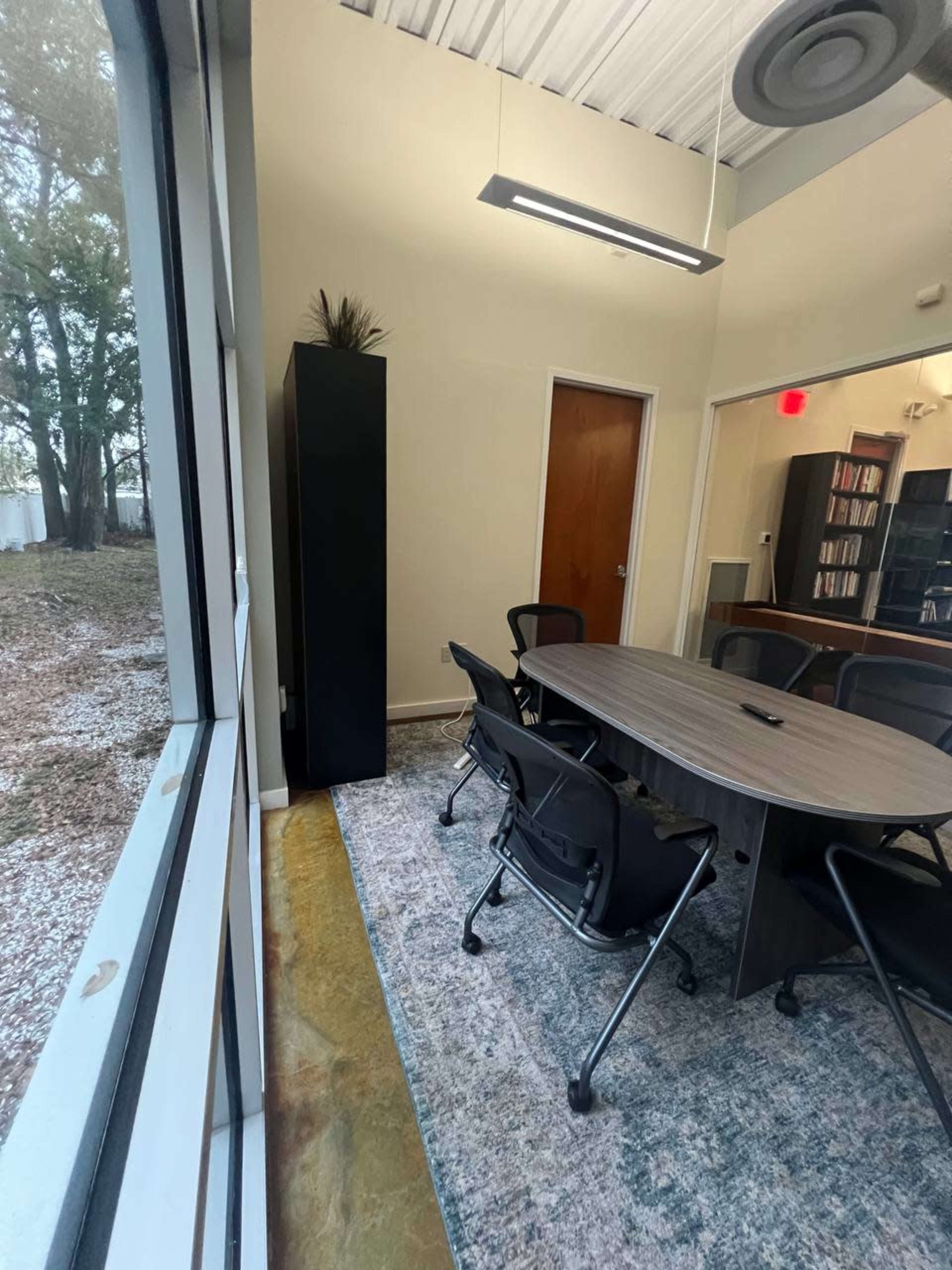 A conference room features a round table surrounded by chairs, with a view of trees through large windows.
