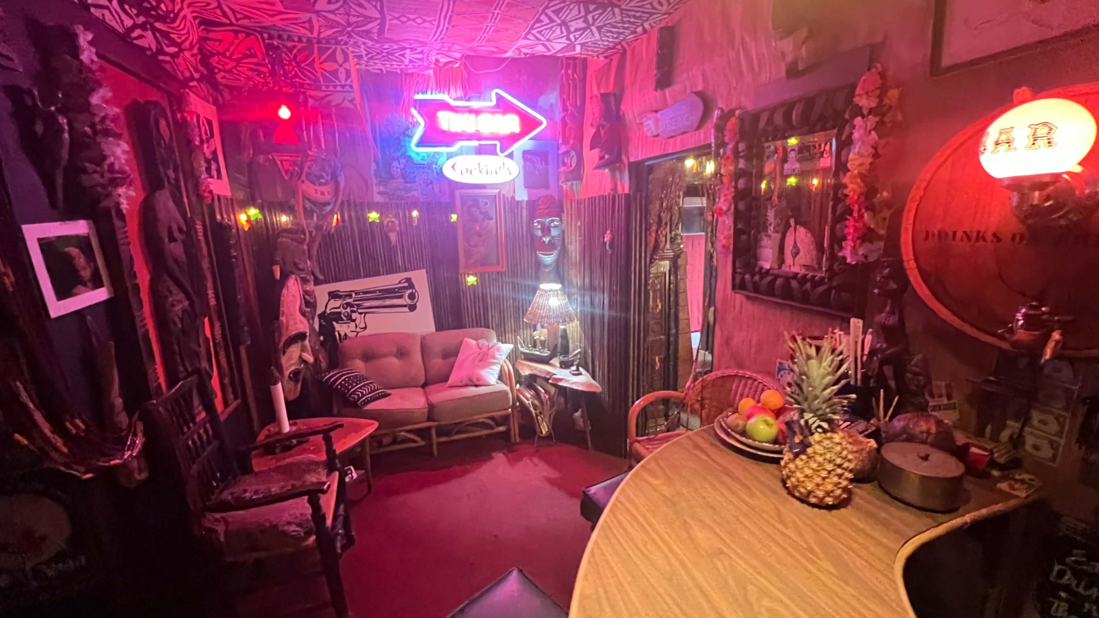 The image shows a dimly lit room decorated with various mirrors, masks, and colorful neon signs, featuring a small table with fruit and a couch.