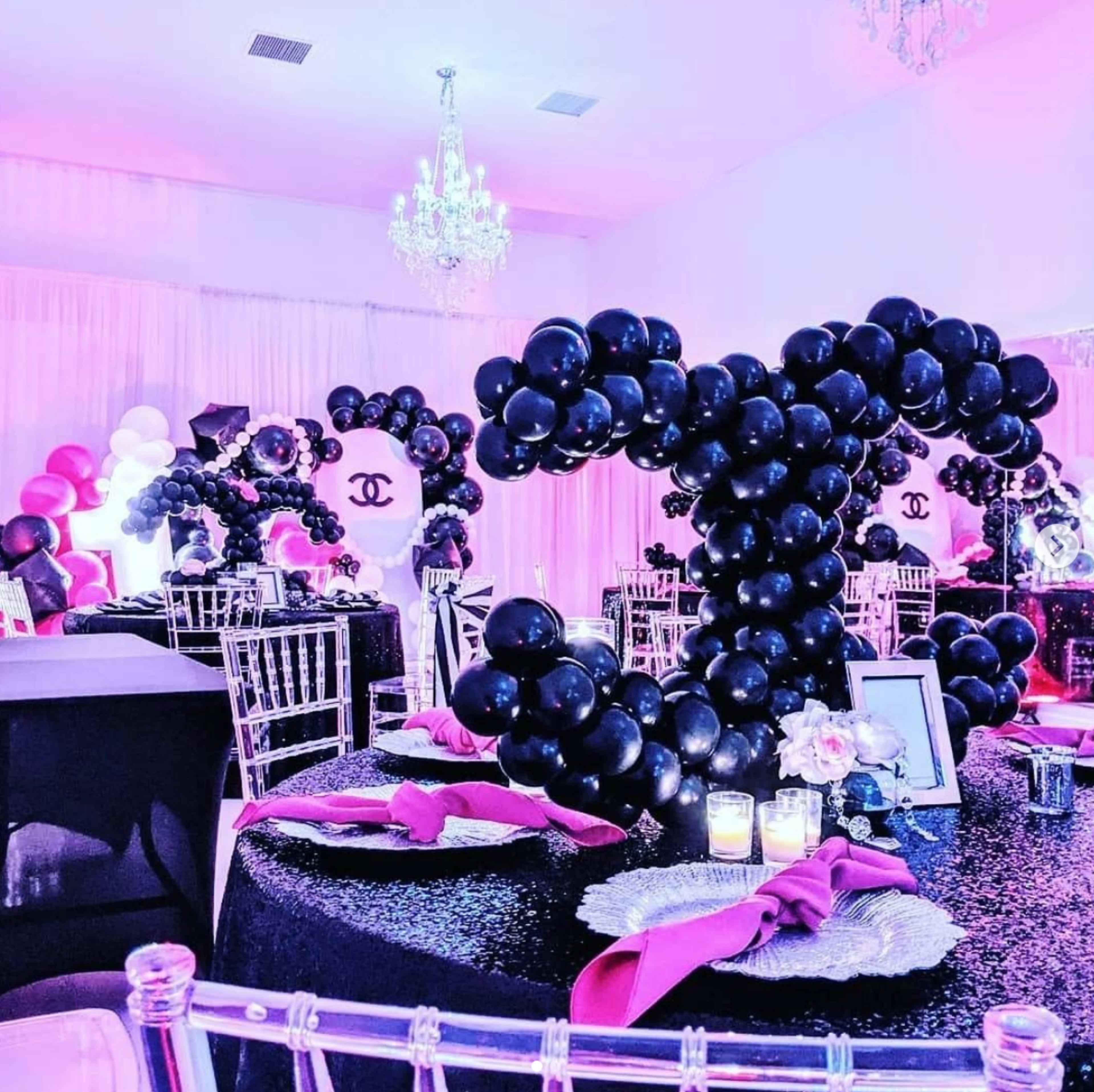 The image shows a decorated event space featuring elegant tables adorned with black and pink balloons shaped like a logo, sparkling tablecloths, and candle centerpieces.