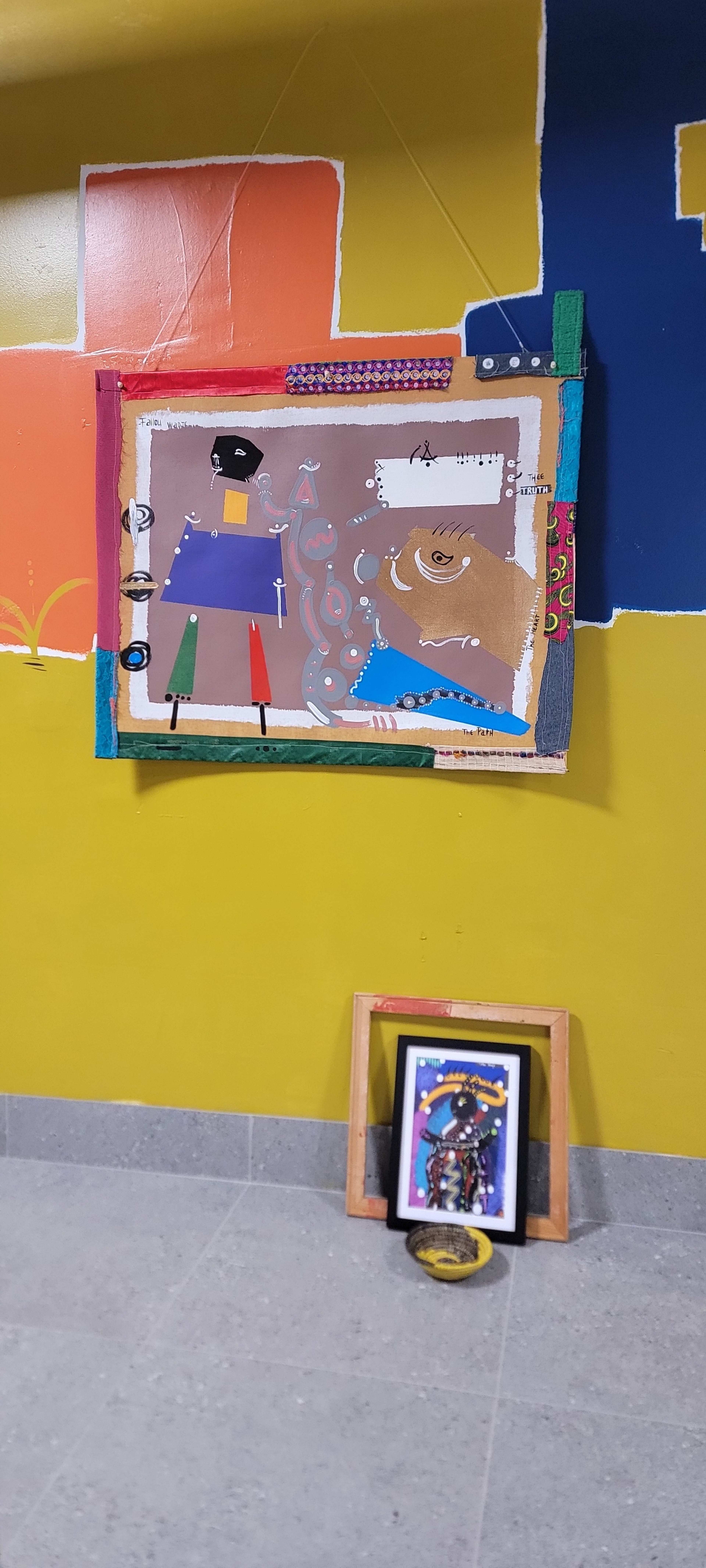 The image displays a colorful wall featuring an abstract painting mounted above a framed artwork positioned on the floor.