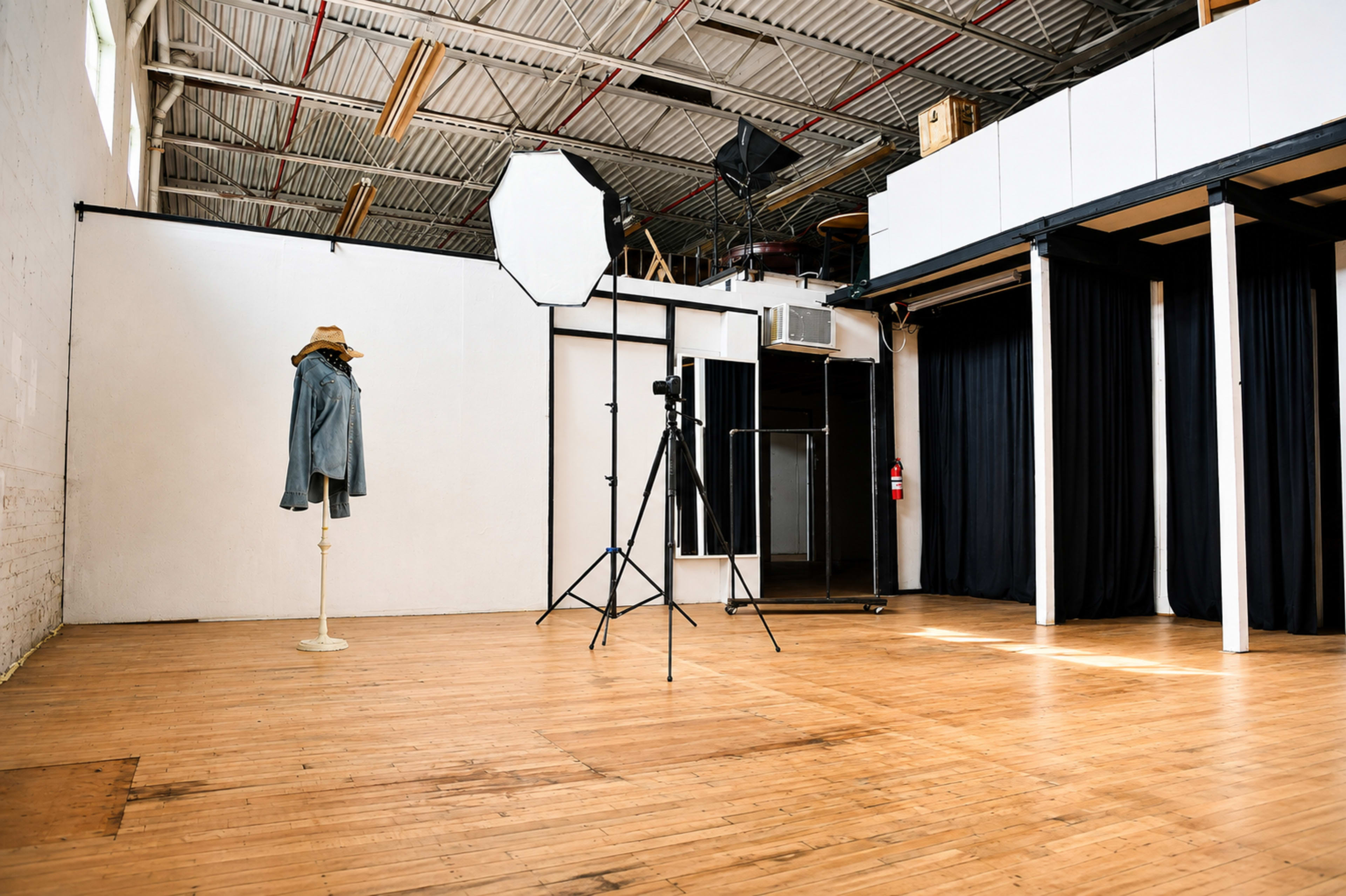 The image shows a spacious photography studio with wooden flooring, featuring a mannequin wearing a jacket, a light umbrella, and a camera on a tripod against a backdrop of black curtains.