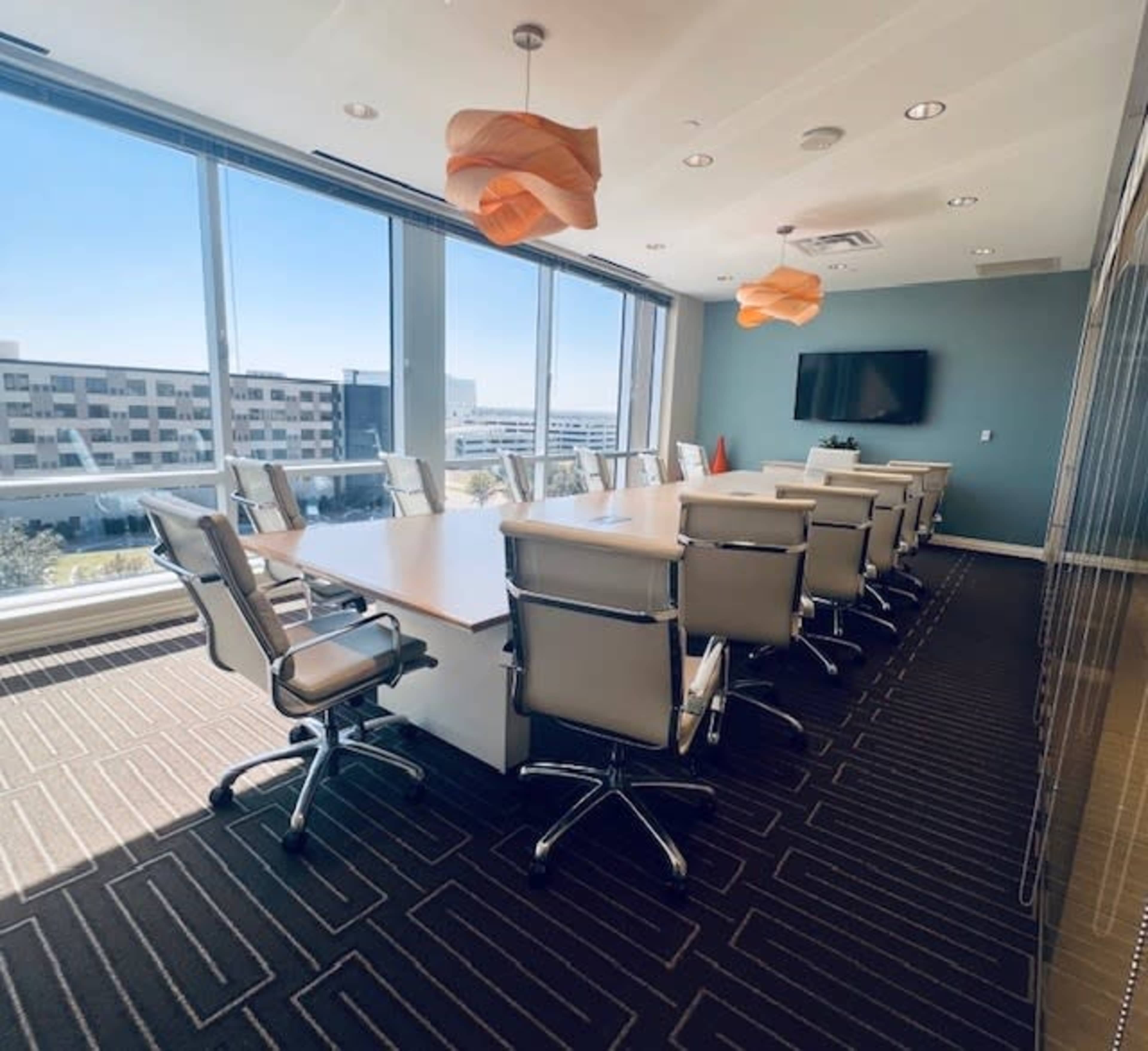 The image shows a modern conference room with a long table surrounded by ergonomic chairs, large windows providing natural light, and a wall-mounted screen.