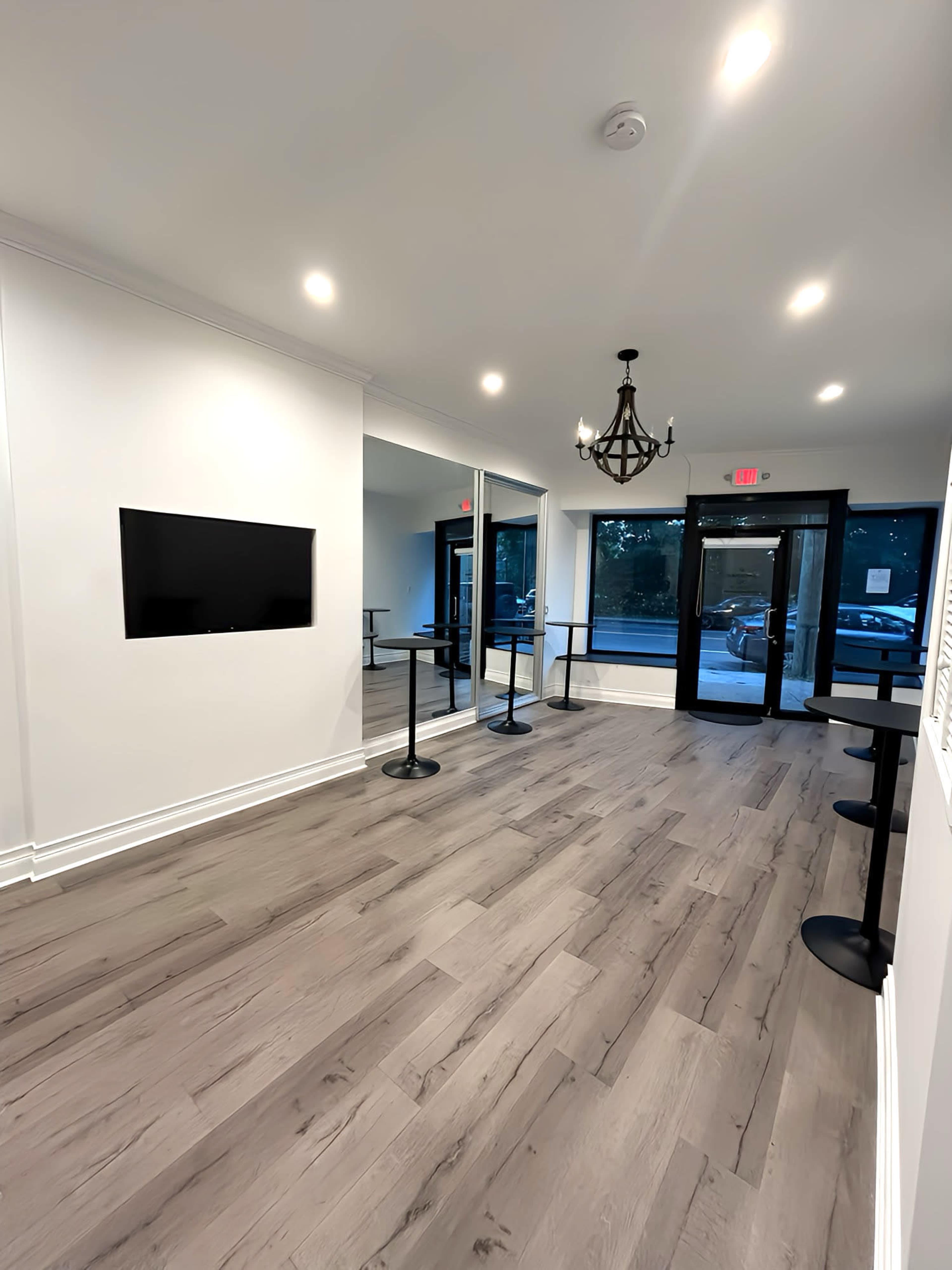 The image shows a modern, well-lit reception area with a TV mounted on the wall and a row of standing tables along the sides, leading to glass double doors that open to an outdoor space.