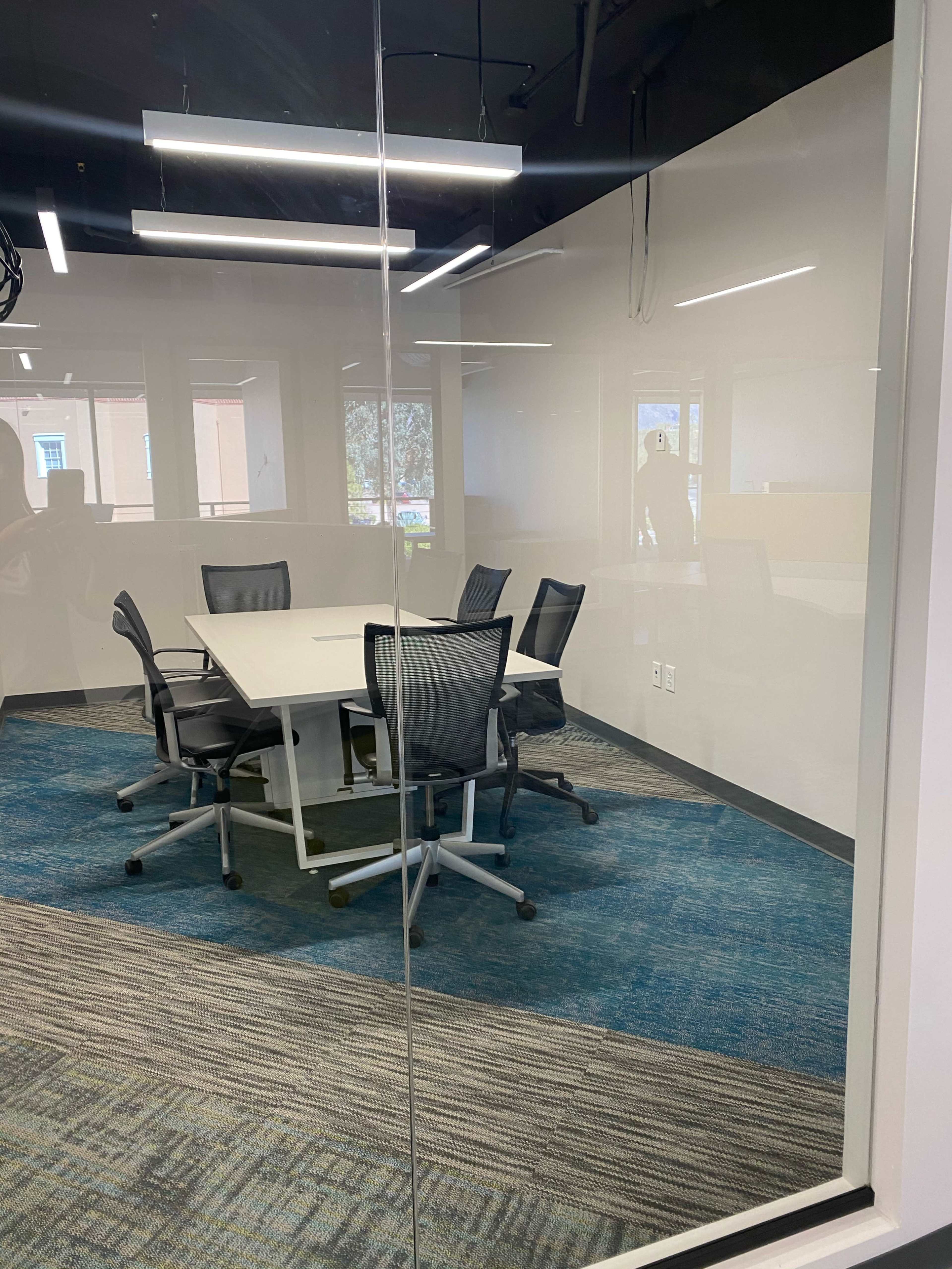 A glass-walled conference room features a round table surrounded by six office chairs on a patterned carpet.