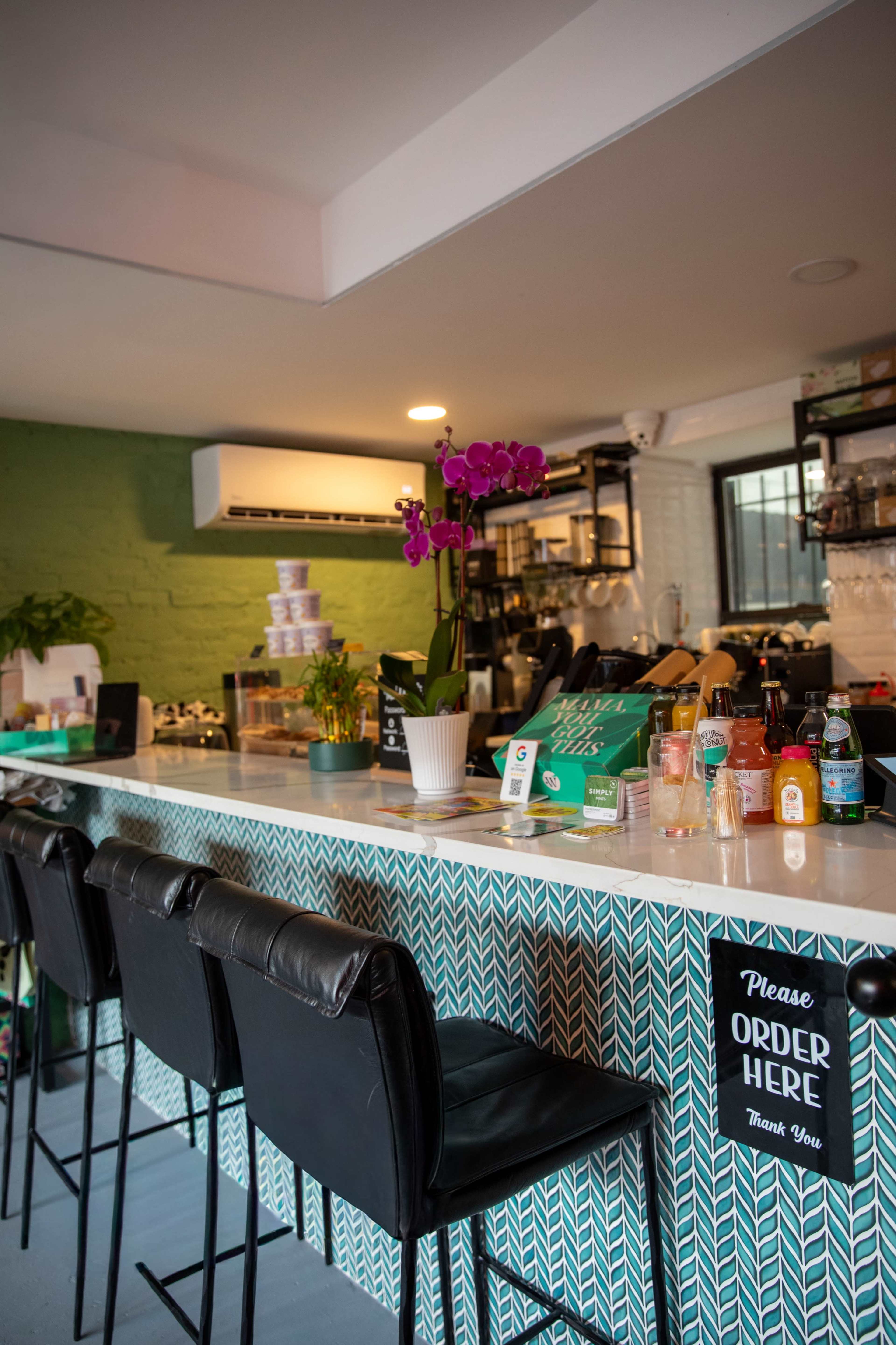 The image shows a café counter with a green patterned backsplash, bar stools, and various beverages and plants arranged on the countertop.