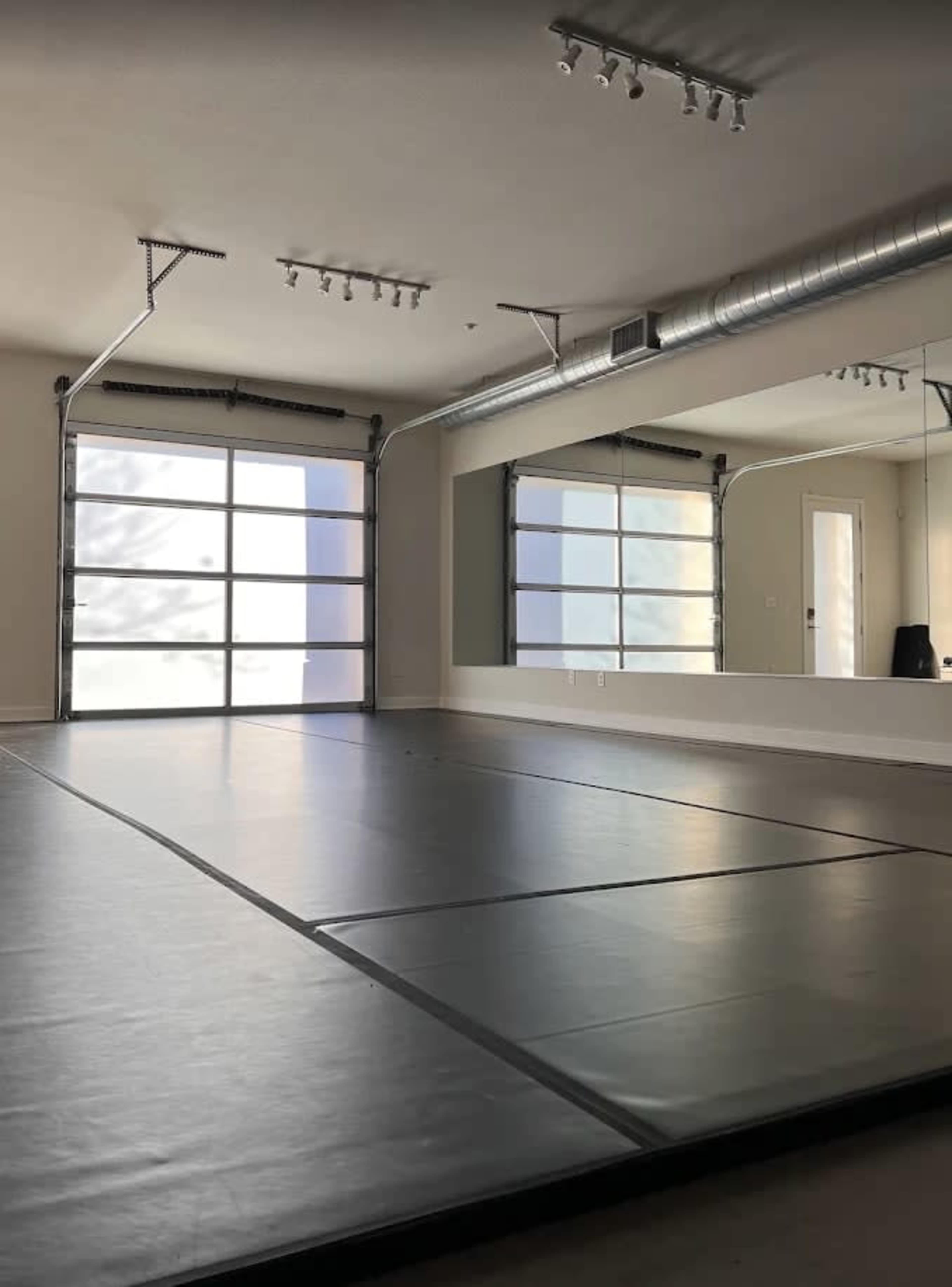 The image shows an empty indoor space with a large mirrored wall and a rolled-out mat on the floor, illuminated by natural light coming through a large window.