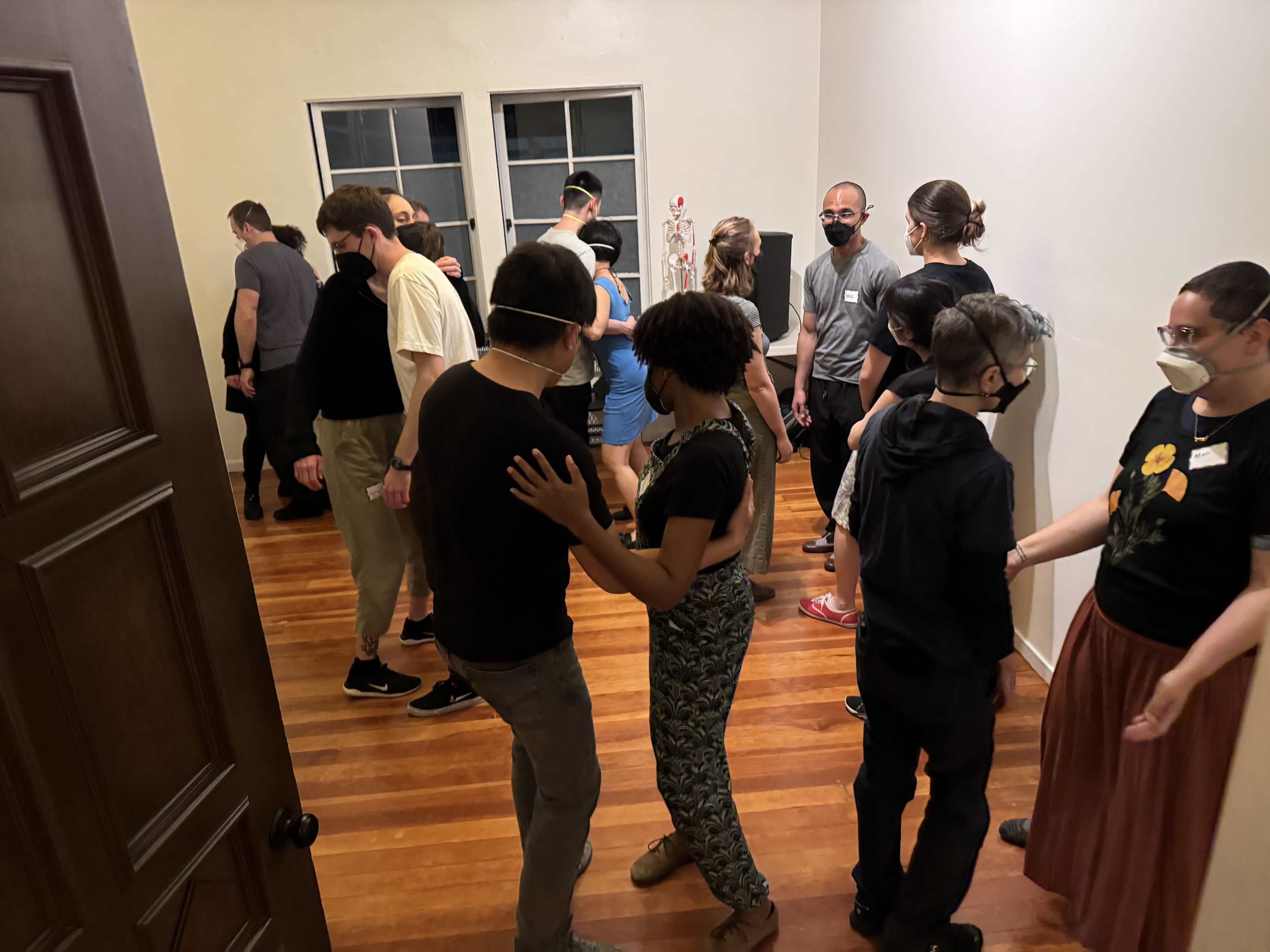 A group of individuals wearing masks participate in a social activity in a room with wooden flooring and white walls.