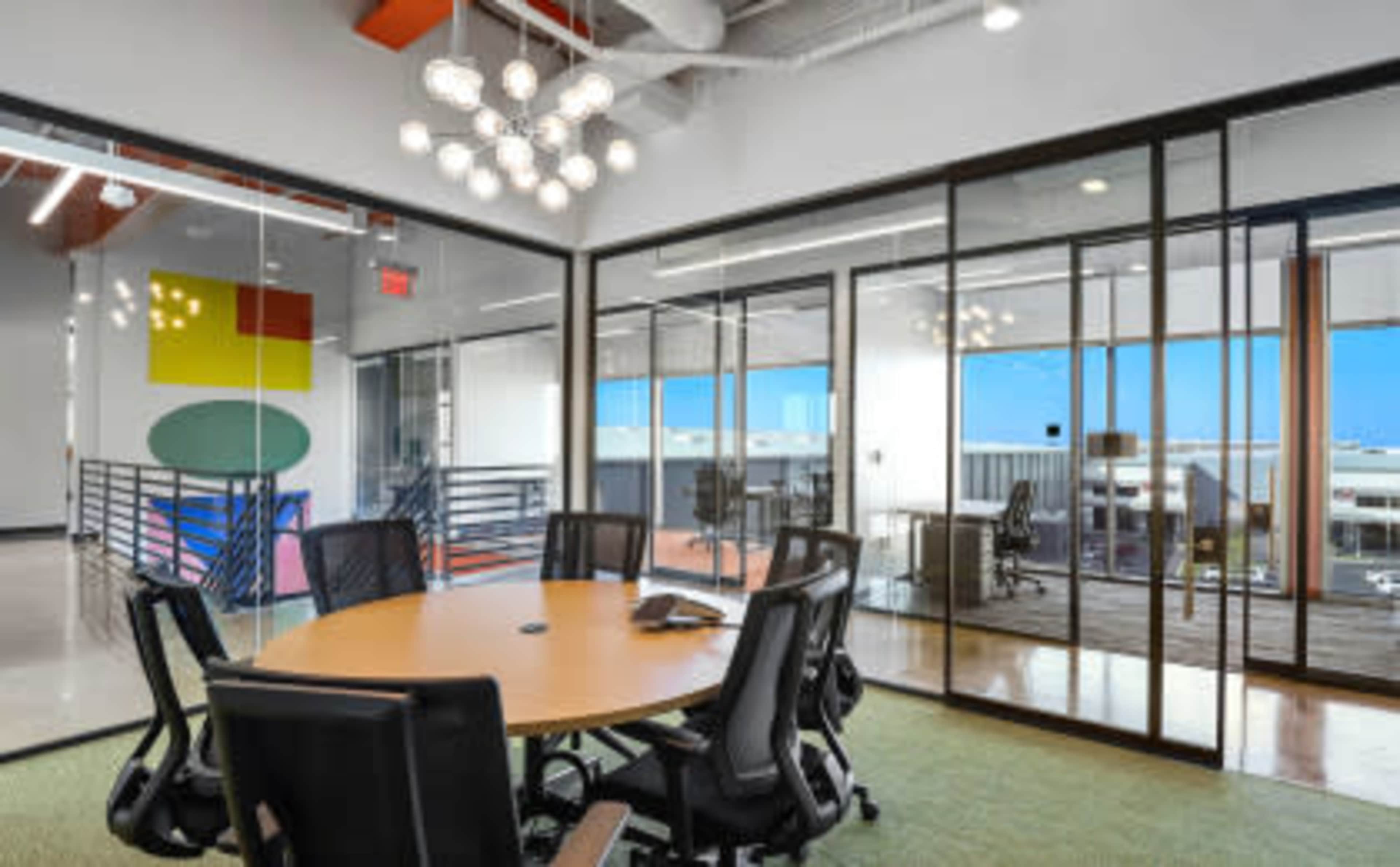 A modern conference room features a round table surrounded by black office chairs, with glass walls providing a view of adjacent work areas.