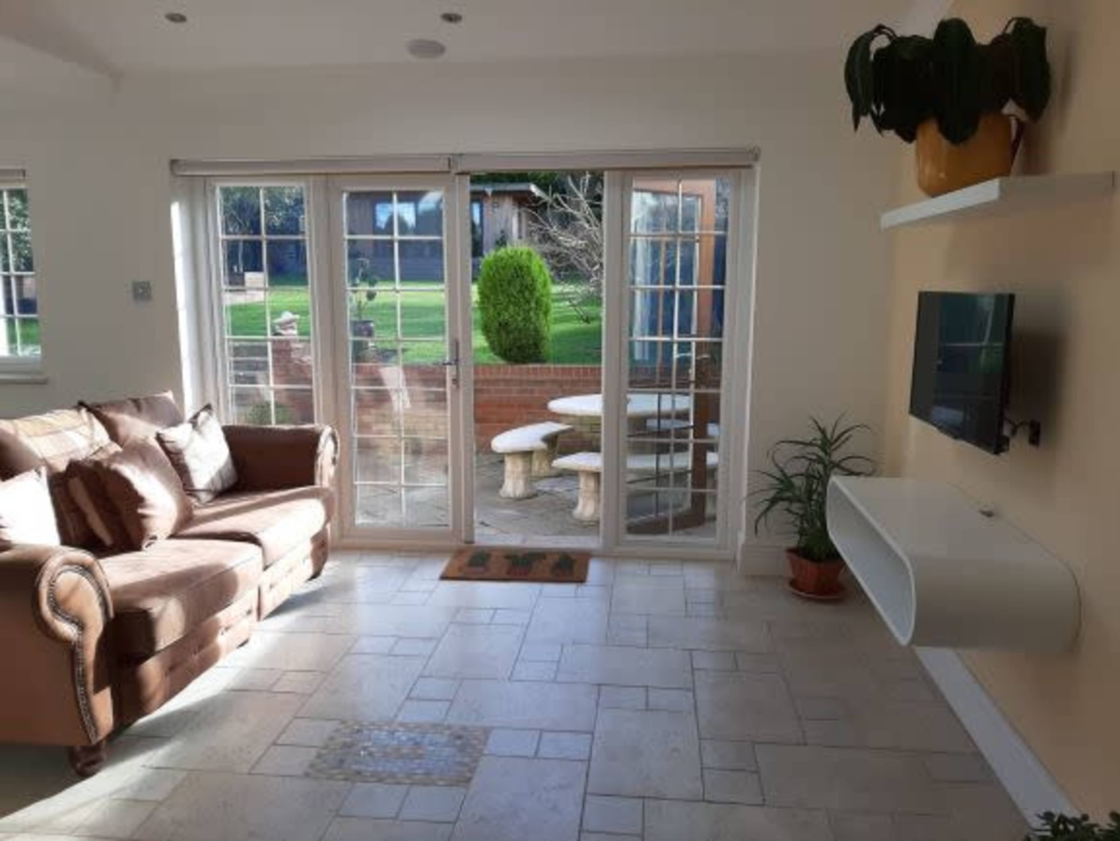 The living room features a brown sofa, a wall-mounted TV, and large glass doors leading to a garden area with a stone table and benches.