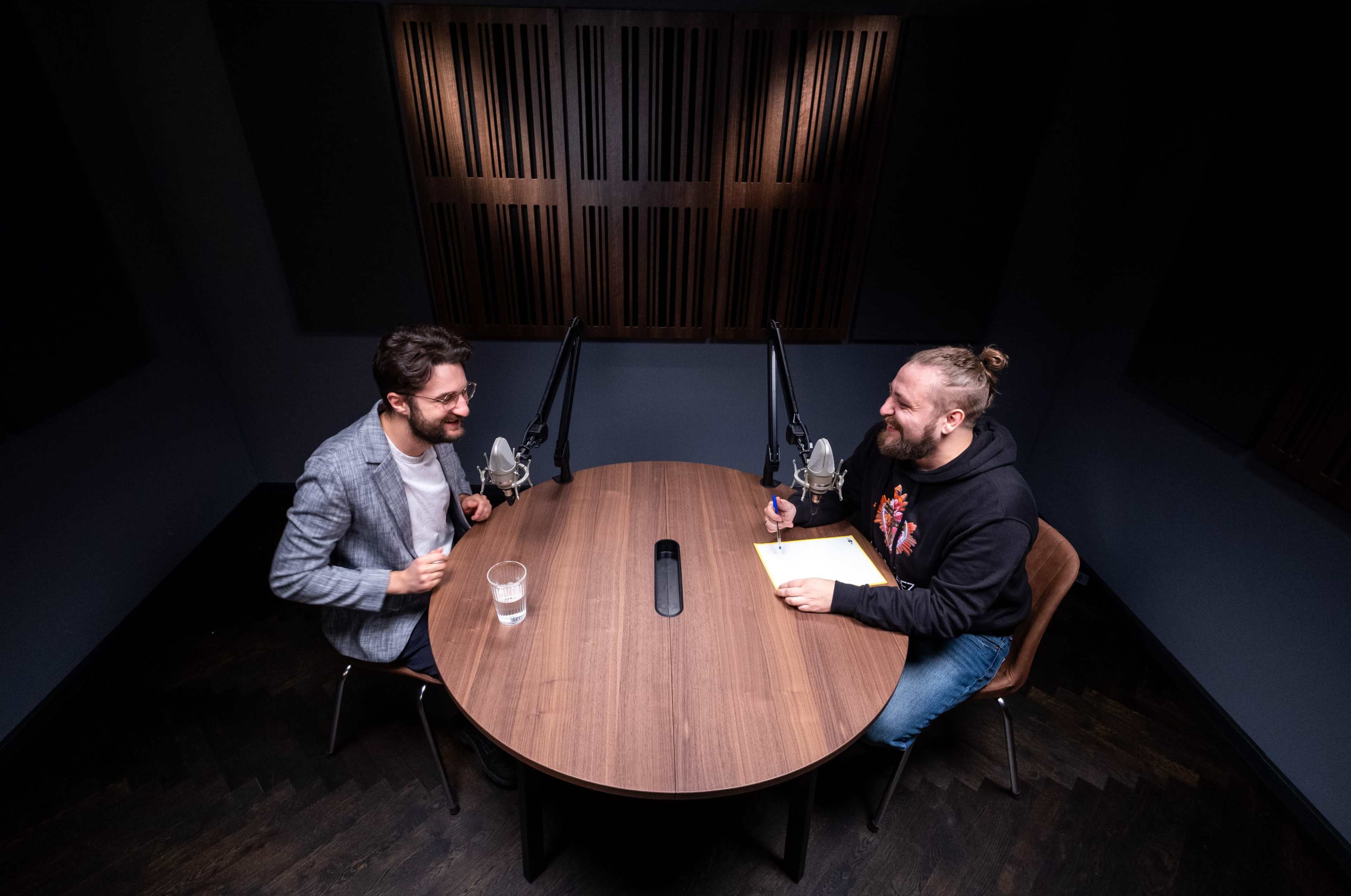 Two men sit at a round wooden table in a recording studio, each facing a microphone, with one holding a notepad and the other a glass of water.