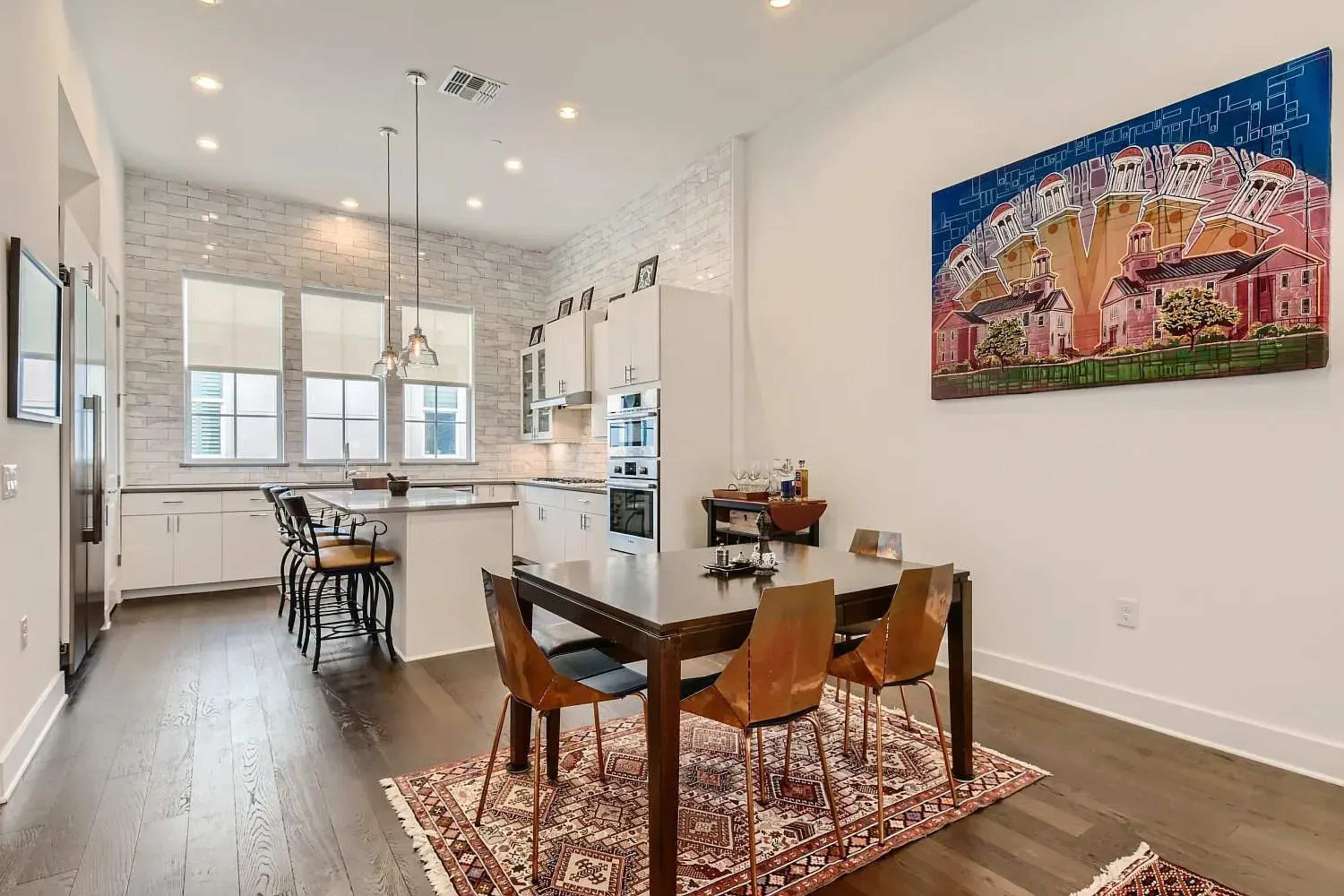A modern kitchen and dining area features a wooden table surrounded by four chairs, with white cabinetry and a colorful painting on the wall.