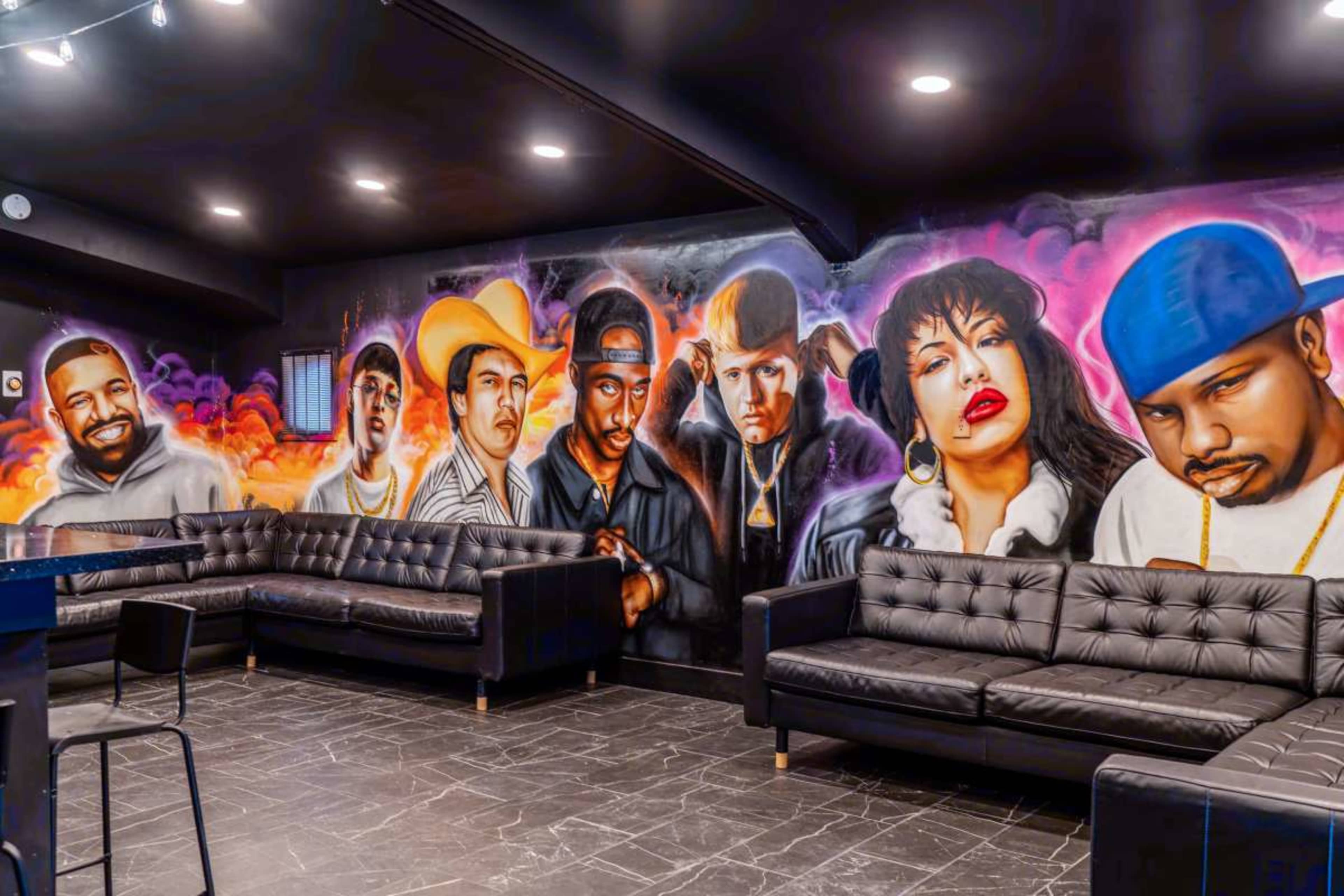 The image shows a mural featuring prominent musical figures on a wall in a lounge area with black leather seating.