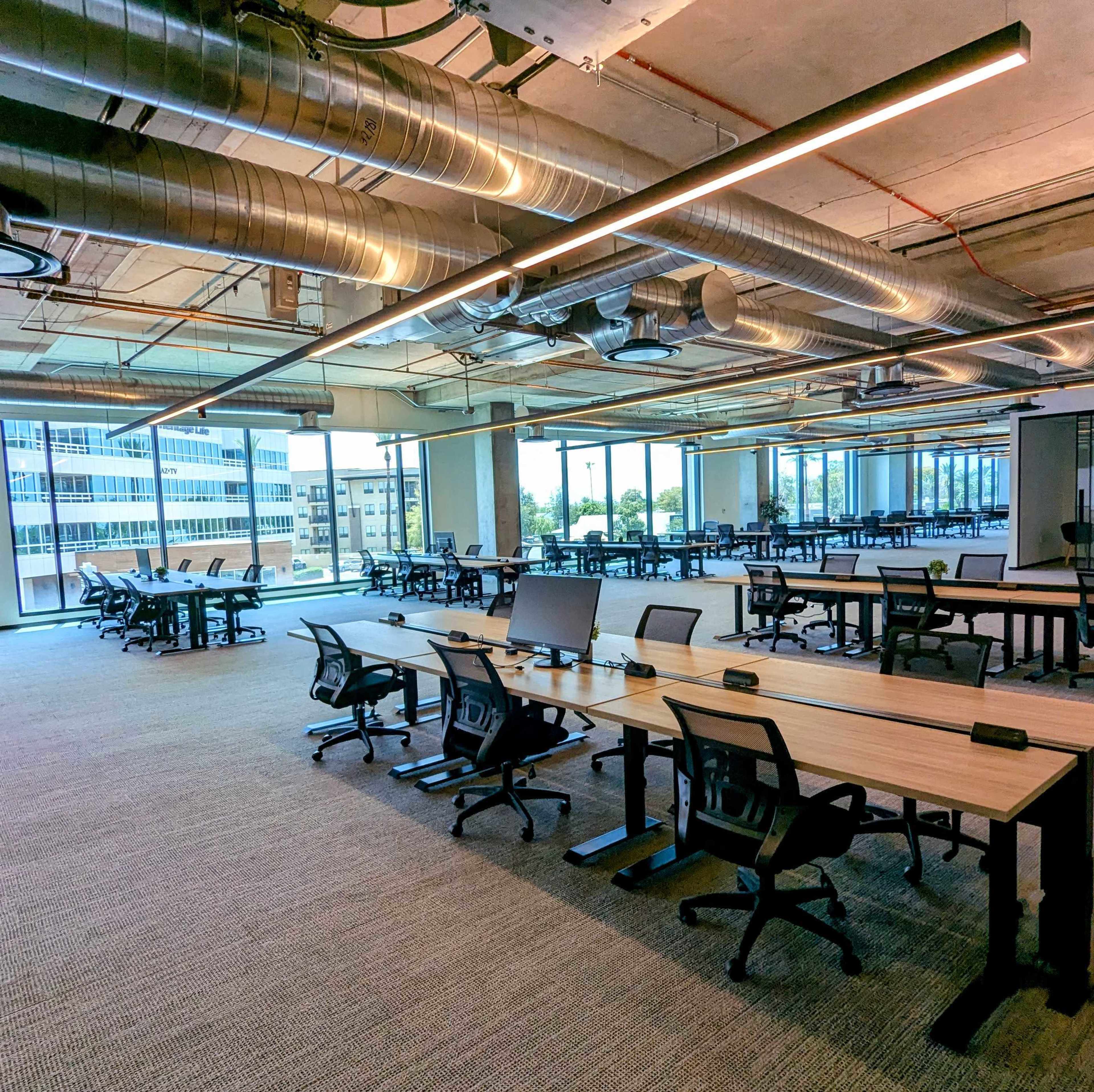 A modern office space with empty workstations arranged neatly under industrial-style lighting and exposed ductwork.