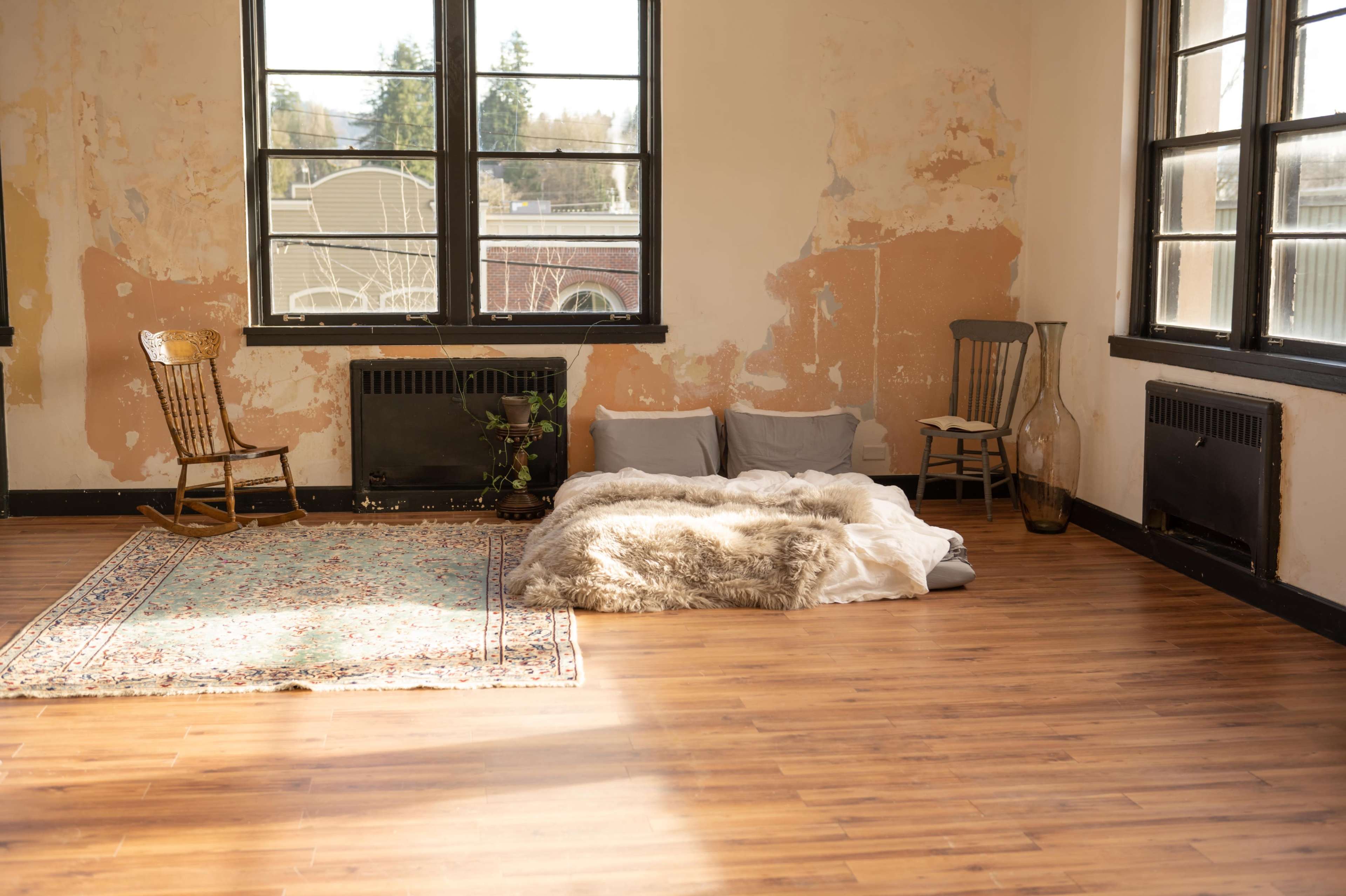 A spacious room with a wooden floor, a worn area rug, and a makeshift bed made of blankets and cushions against a wall with peeling paint.