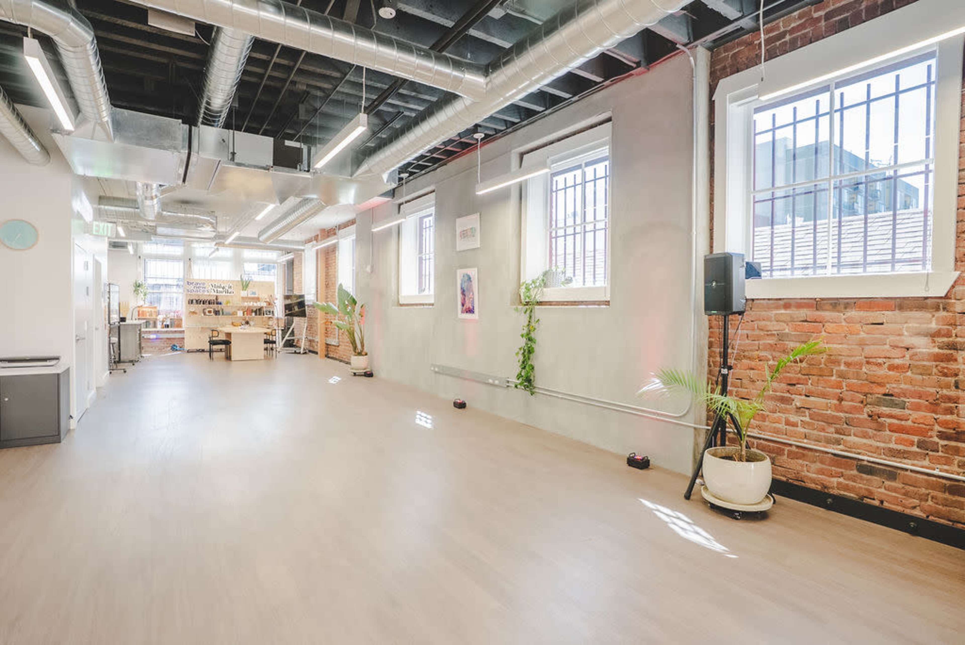 The image depicts a spacious, modern interior with exposed brick walls, large windows, and wooden flooring, featuring plants and an open area that appears to be a multipurpose space.