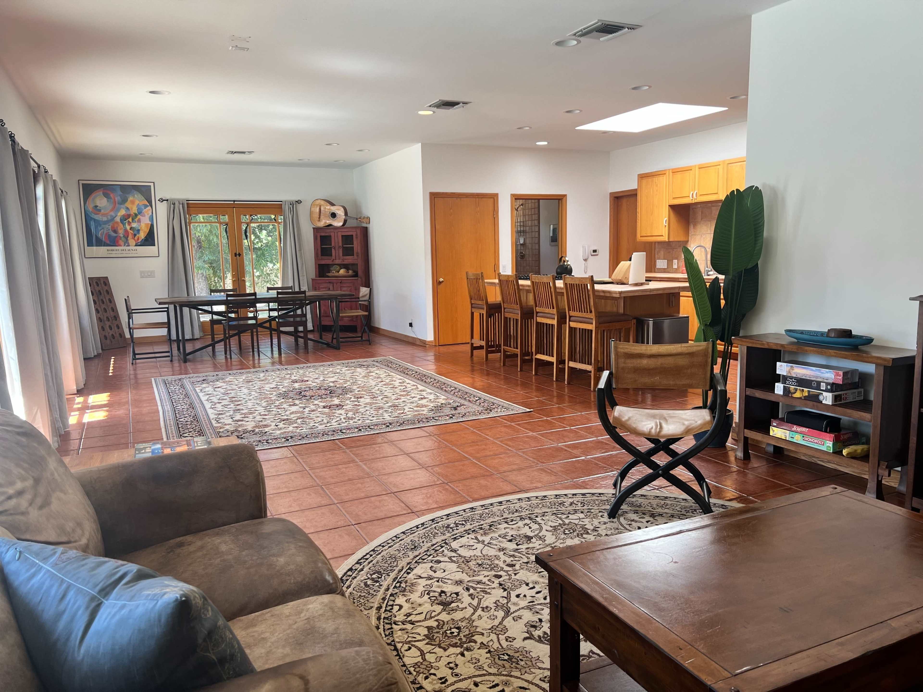 The image depicts a spacious living area with a seating arrangement, dining table, and kitchen, featuring tiled floors and natural light from large windows.