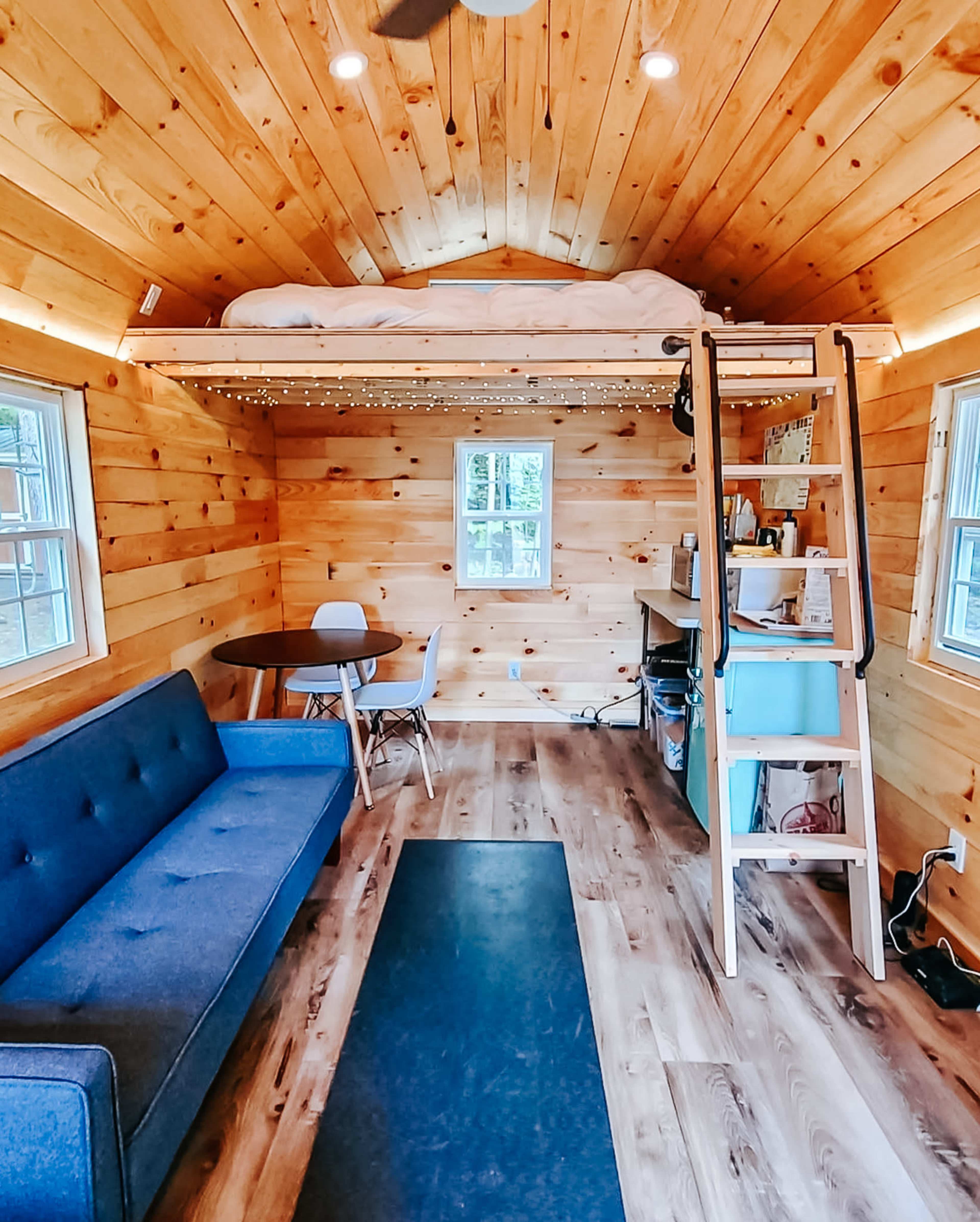 The interior of a tiny house features a lofted bed, a gray sofa, a round table with chairs, and a ladder leading to the sleeping area, all surrounded by wood-panelled walls.
