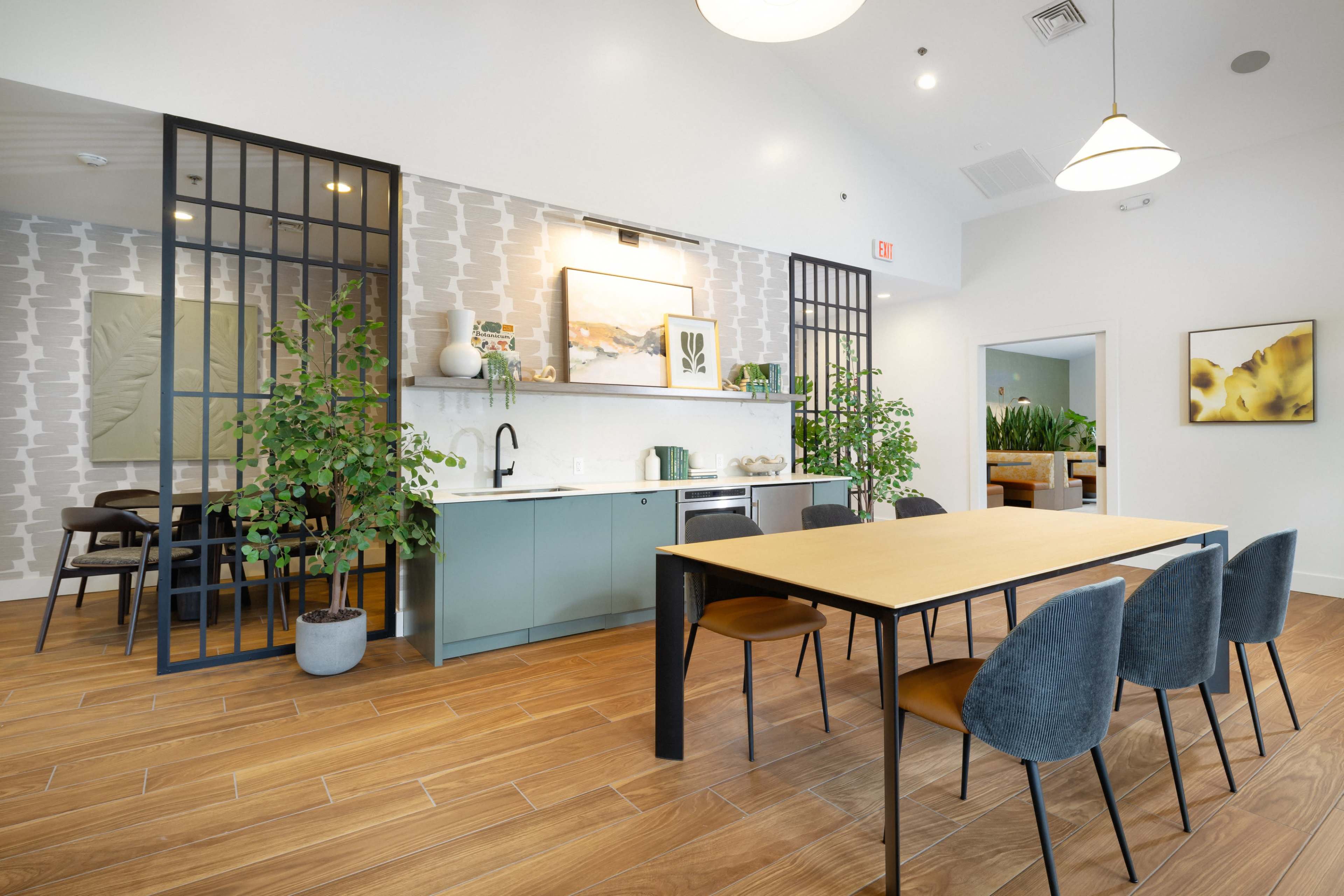The image shows a modern dining area with a large table surrounded by chairs, a kitchenette along one wall, and decorative plants.