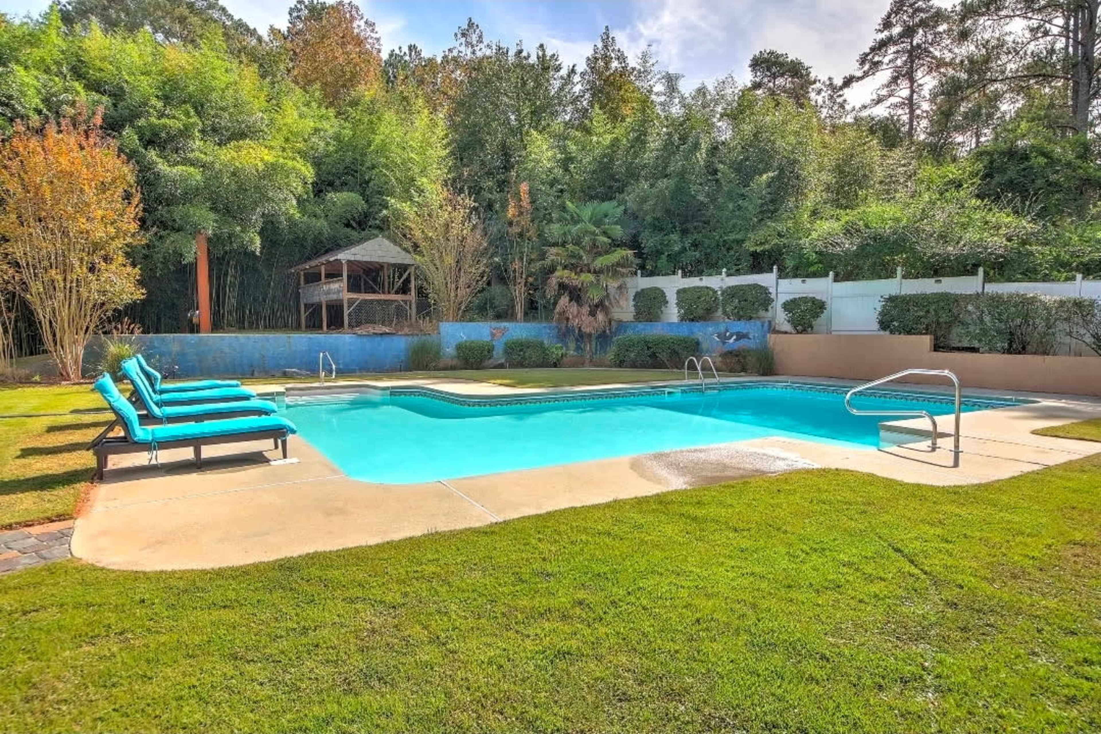 The image shows a swimming pool surrounded by green grass, with several blue lounge chairs set up nearby and a gazebo in the background.
