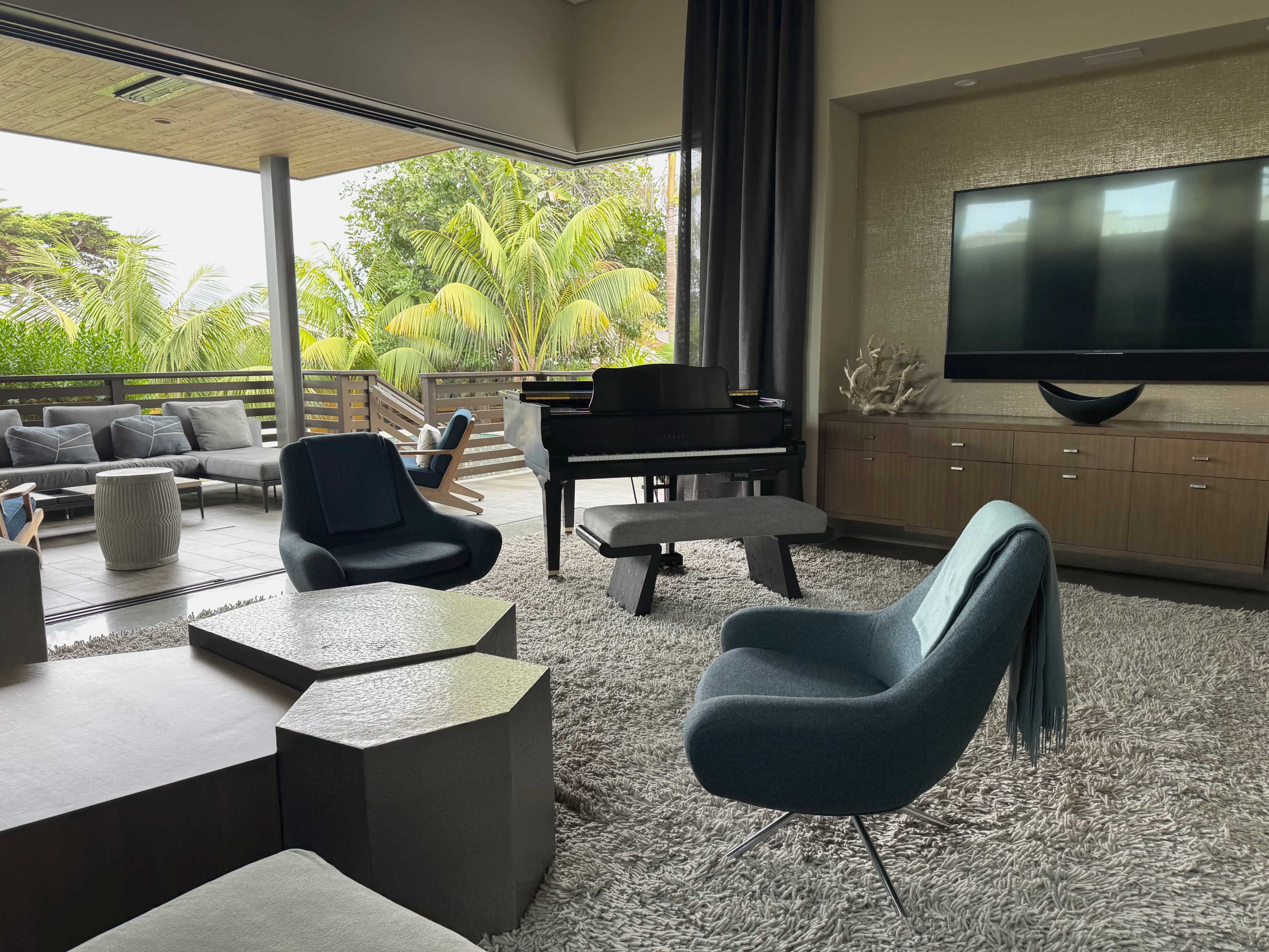 The image shows a modern living room with a grand piano, sleek furniture, and large windows that open to a patio with tropical plants.